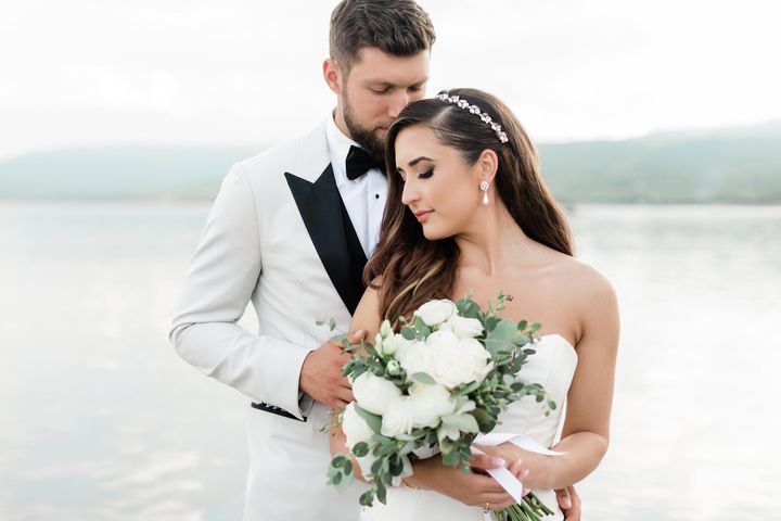A bride and groom in white standing together; the bride wearing a crystal bridal headband, pearl bridal earrings, and a white bridal bouquet with ocean background