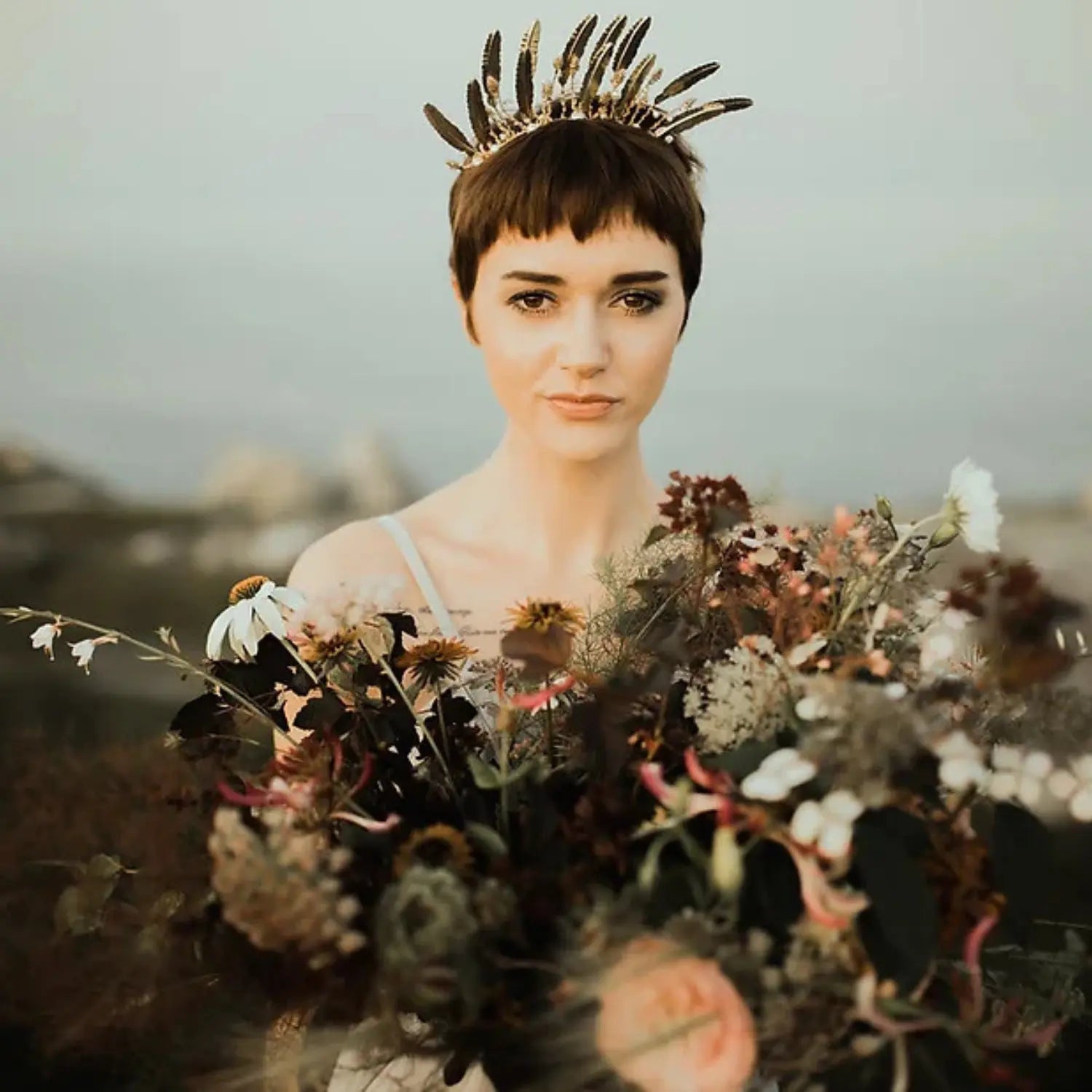 A woman holding a bouquet of wild flowers while wearing a golden feather crown.