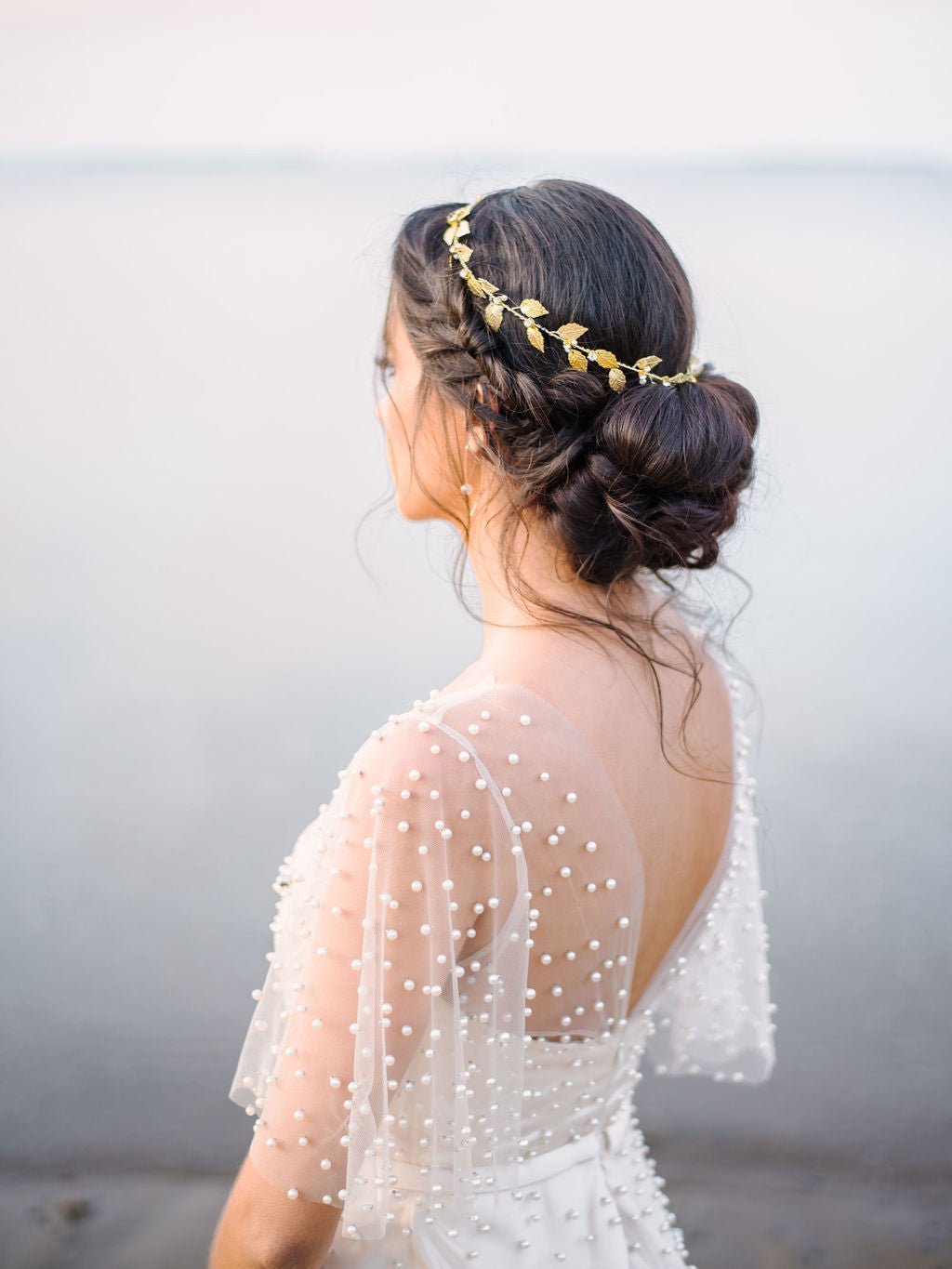 Bride in white pearl bridal gown displaying a beautiful golden bridal hair circlet with dainty freshwater pearls and crystals among glistening leaves on bridal up-do with grey background. Pearl bridal accessories, gold bridal accessories, & bridal hair accessories Canada