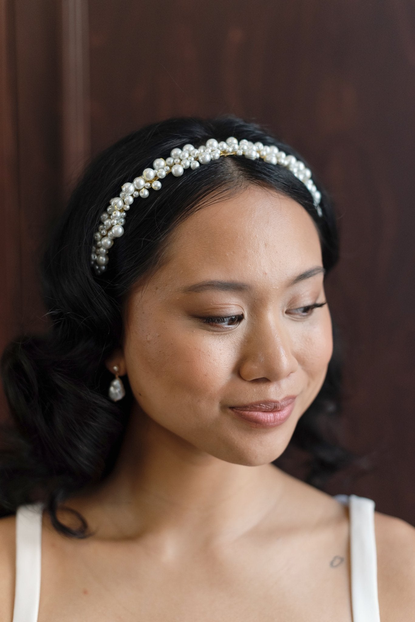 Smirking BIPOC bride wearing a golden bridal headband with large Swarovski pearl clusters on dark wooden background. Bridal hair accessories & Handmade Canadian jewelry by BIPOC designer