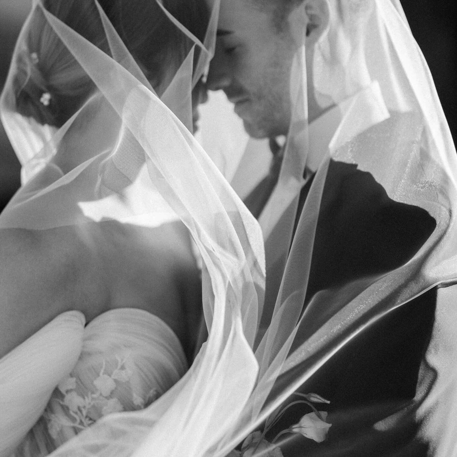 A black and white photo of a newlywed couple touching foreheads shrouded in a sheer tulle wedding veil