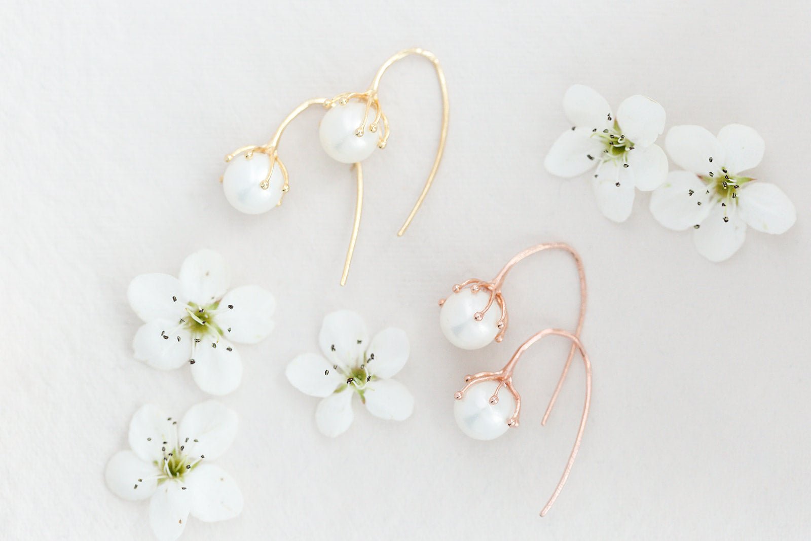 Two pairs of Bloom bud and freshwater pearl earrings in rose gold and gold-filled with white flowers on neutral background. Pearl Bridal jewelry & Organic pearl jewelry handmade by Canadian jewelry artisan