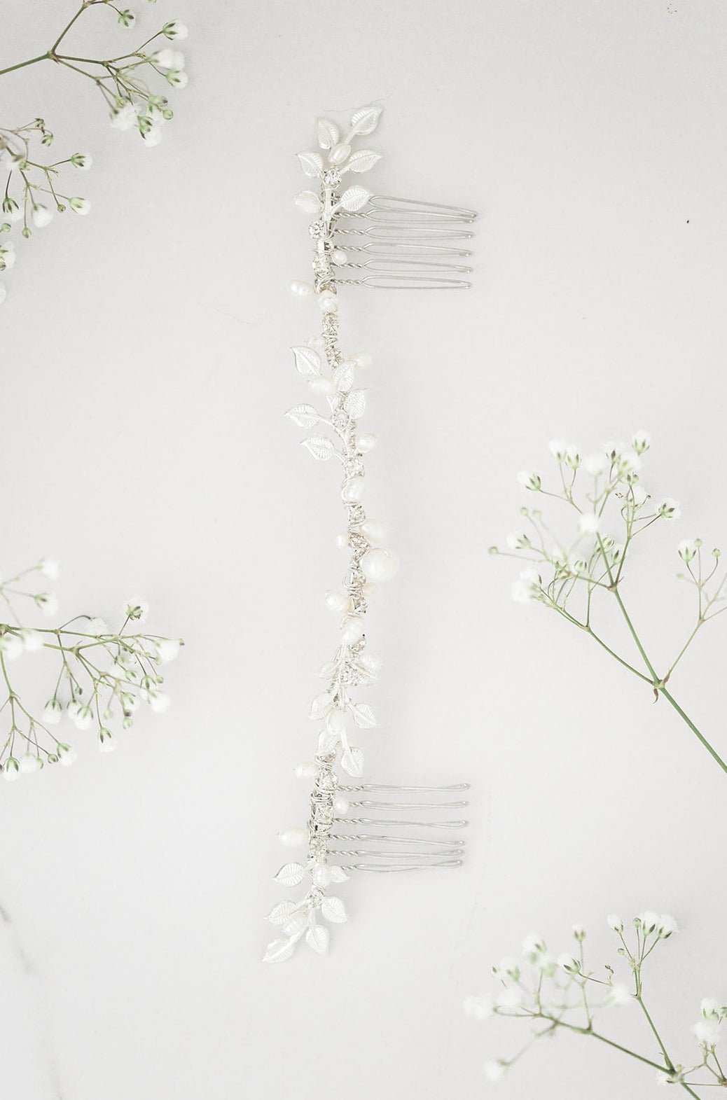 A dainty Silver Byrony Fresh Water Pearl, Crystal, and Leaf Hairvine is placed amongst some baby's breath. Bridal hair accessories, pearl bridal accessories, & affordable bridal accessories handmade by small Canadian jewelry brand
