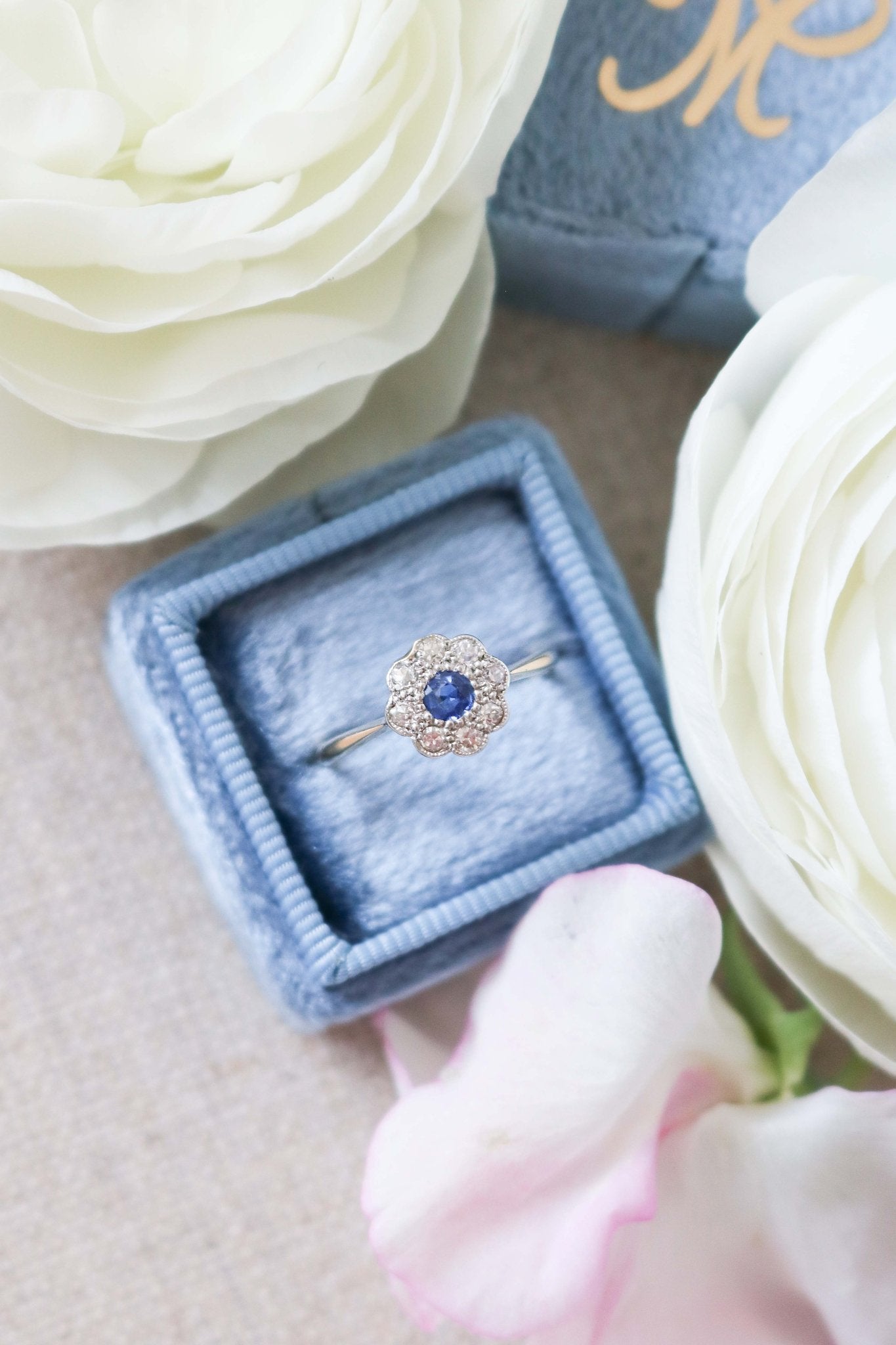Daphne 18k Yellow and White Gold Vintage Sapphire and Diamond Engagement Ring in a flower design sits amongst roses in a blue ring box. Canadian vintage engagement rings. Bridal jewelry Canada. Canadian weddings. Wedding Canada. Canadian bridal jewelry. Woman-owned Canadian jewelry brand. Canadian jewelry brands.
