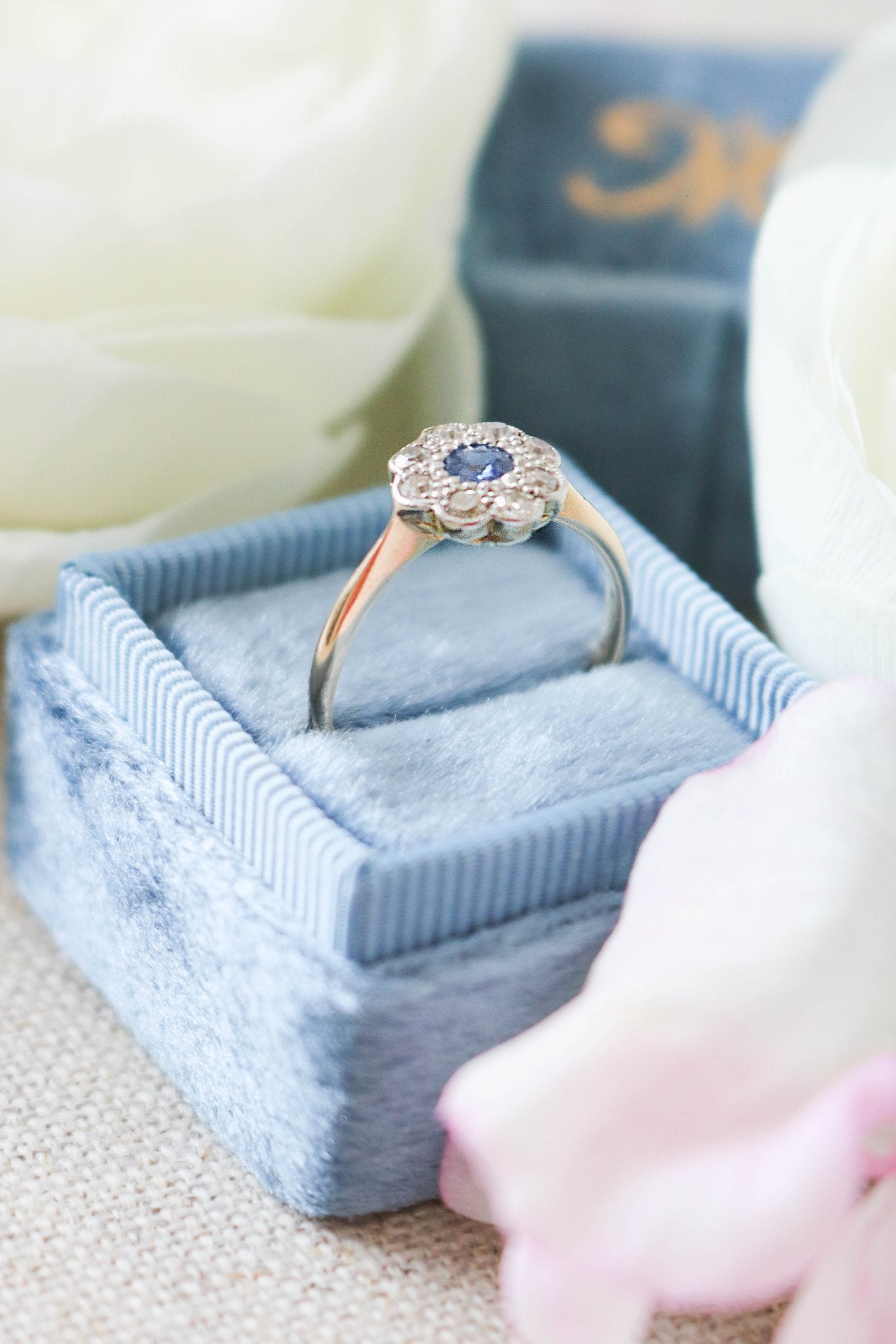 A close up side view of an 8k Yellow and White Gold Vintage Engagement Ring encrusted with a sapphire and multiple diamonds sitting in a blue ring box with white roses. Canadian vintage engagement rings. Bridal jewelry Canada. Canadian weddings. Wedding Canada. Canadian bridal jewelry. Woman-owned Canadian jewelry brand. Canadian jewelry brands.