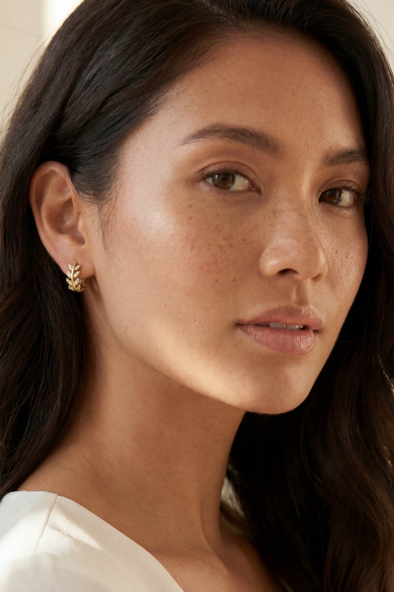 Close-up of BIPOC woman wearing a gold hoop laurel leaf earring with cubic zirconia crystals on a neutral background