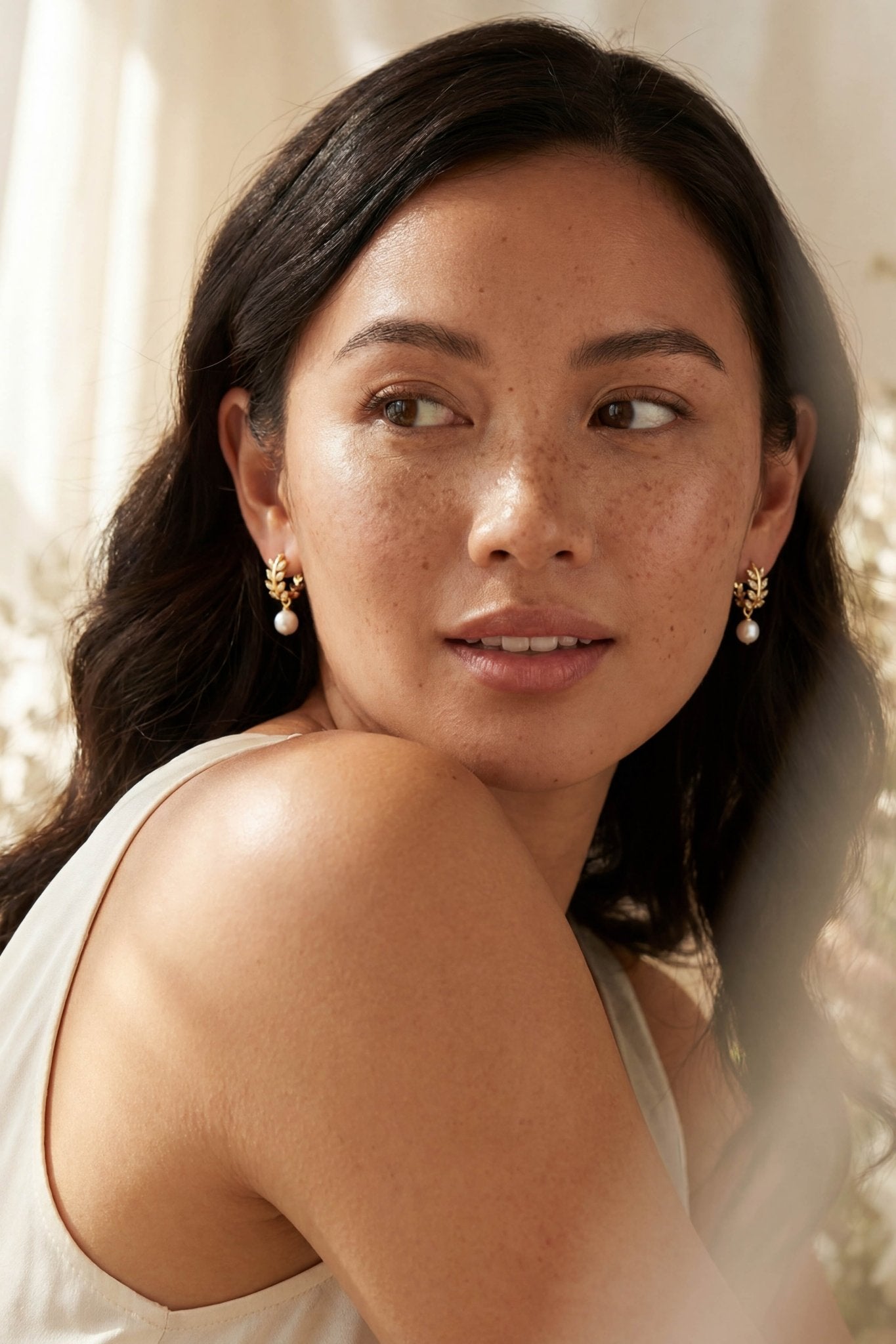 BIPOC woman with long dark hair and gold laurel leaf pearl drop earrings, wearing a white top, against a soft, blurred background. Handmade gold hoop earrings by BIPOC jewellery artist