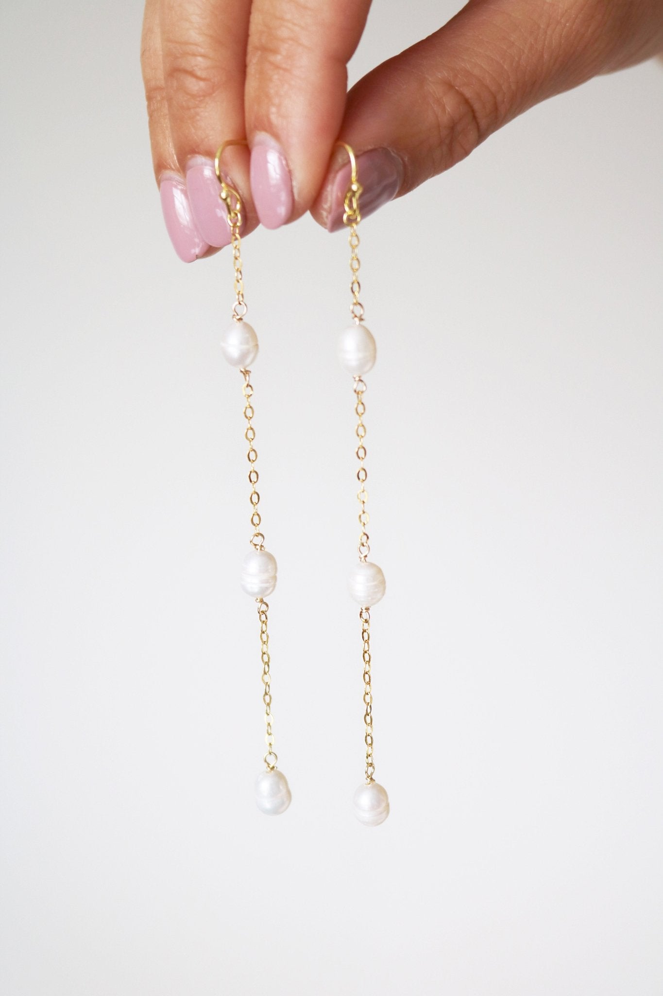 Fingers dangle a pair of drop-down gold-filled freshwater pearl bridal earrings. Organic pearl jewelry. Bridal jewelry Canada. Pearl jewelry Canada. Pearl Bridal jewelry Canada. Gold-filled jewelry Canada. Canadian weddings. Affordable Canadian jewelry brands. Handmade Canadian jewelry.