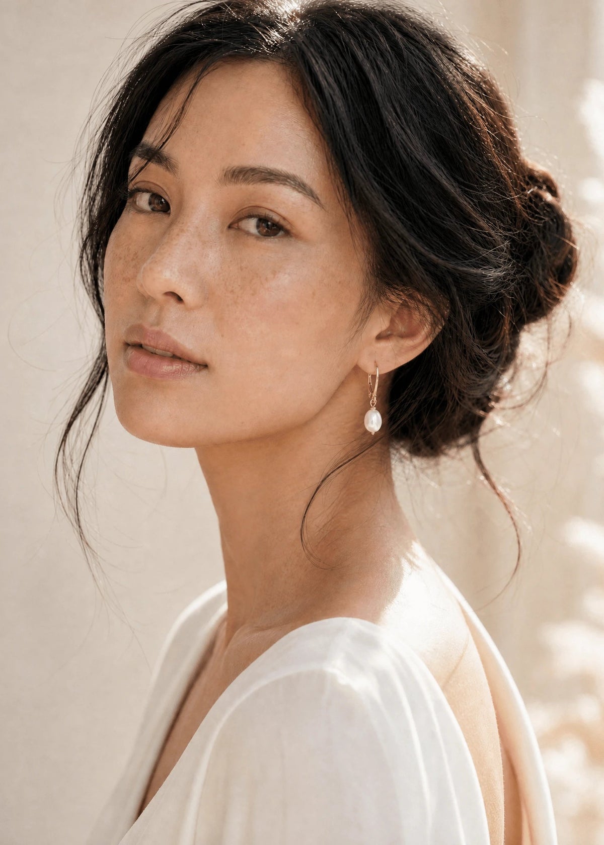 Filipino woman with messy bun in white wearing gold fresh water pearl earrings with leverback hooks against neutral background