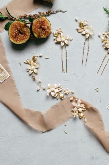 Three bridal hairpins with golden gilded leaves, freshwater pearls, and enamel flowers in a satin pearl hue, displayed with matching bridal hairvine on a pale grey surface with neutral ribbon and halved fig fruit. Pearl bridal hair accessories, affordable bridal accessories, and bridal hair accessories handmade by Canadian bridal designer