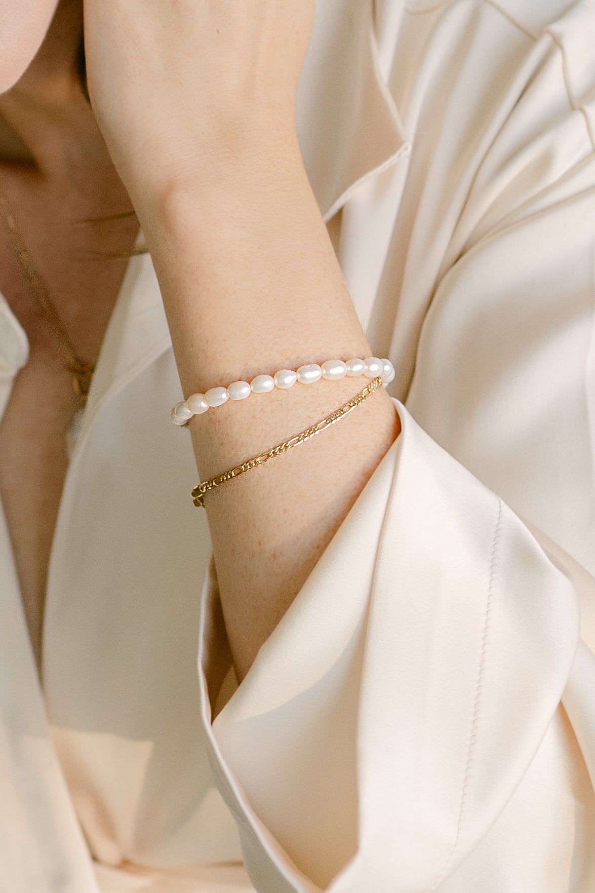 A woman's arm with two bracelets on; an organic freshwater pearl bracelet & gold-filled Figaro Italian style bracelet against a silky off-white shirt. Everyday Jewelry & minimalist jewelry handmade in Canada by Canadian jewelry artisan