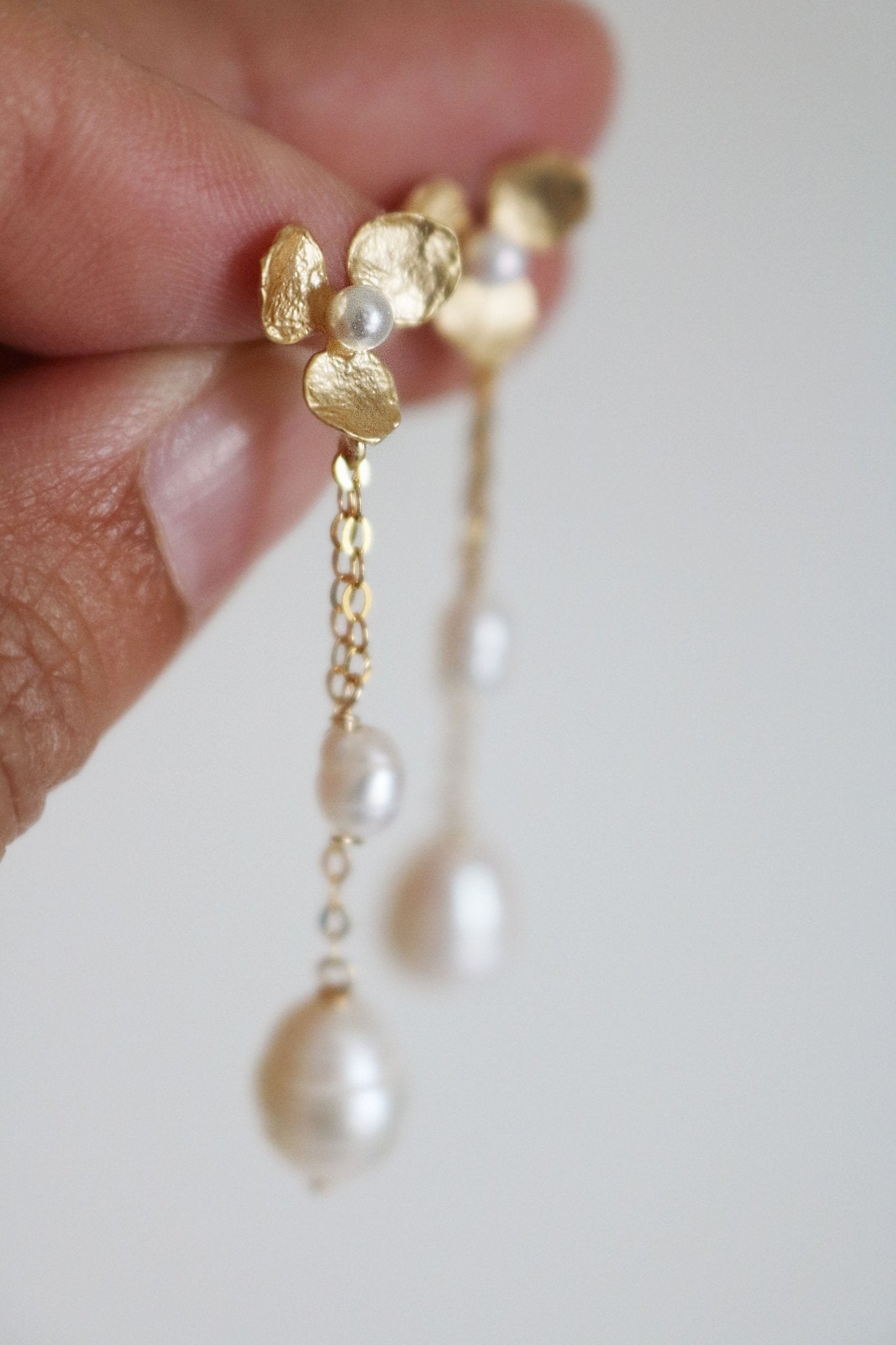 Fingers hold up a pair of elegant floral gold-filled earrings; freshwater pearls dangle down. Bridal jewelry Canada. Pearl jewelry Canada. Pearl Bridal jewelry Canada. Gold-filled jewelry Canada. Handmade Canadian jewelry. Organic pearl jewelry.