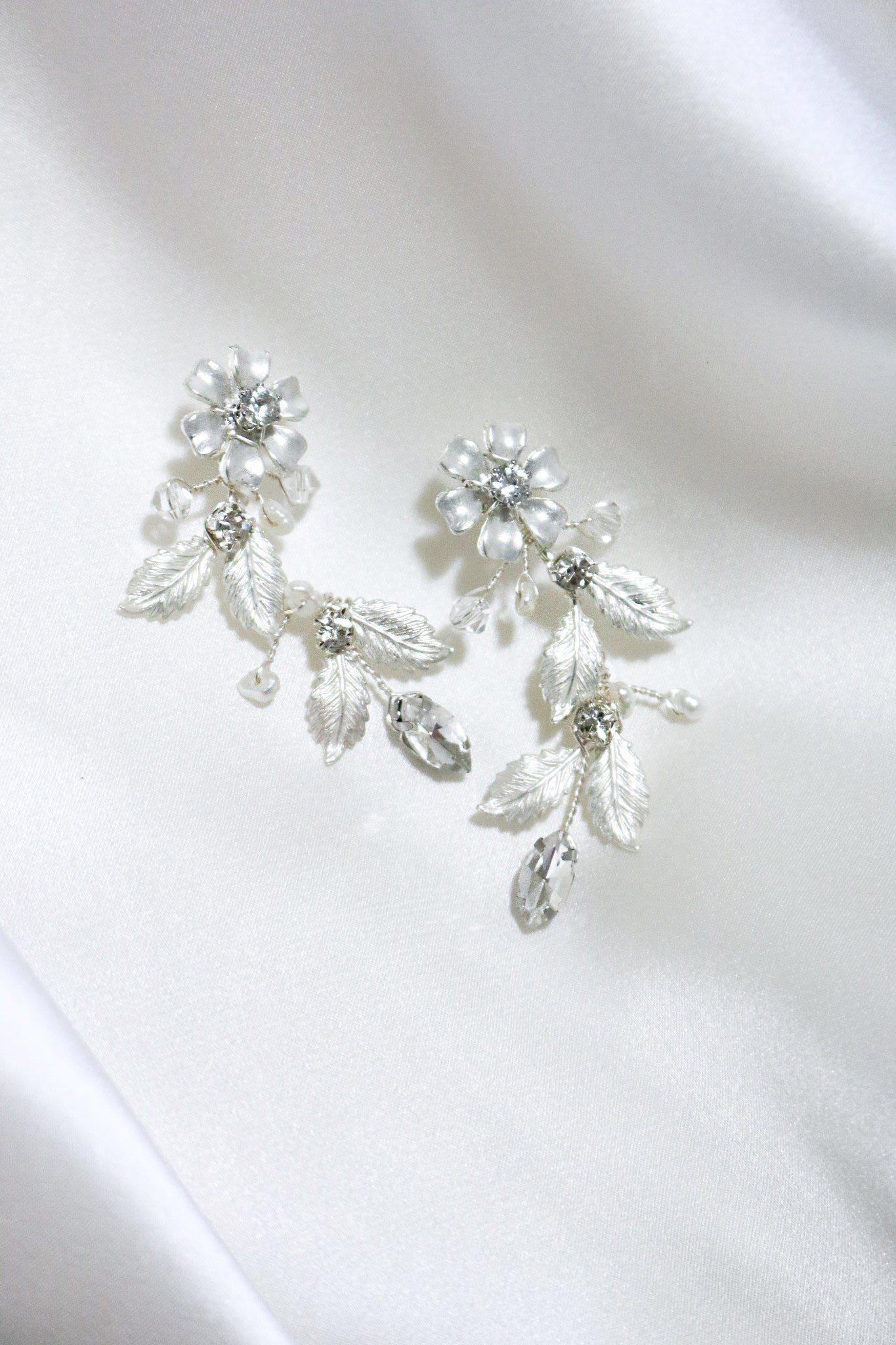 A pair of handcrafted bridal earrings with soft flowering petals, adorned with Swarovski crystals on a white fabric surface. Statement bridal earrings & Bridal jewelry Canada