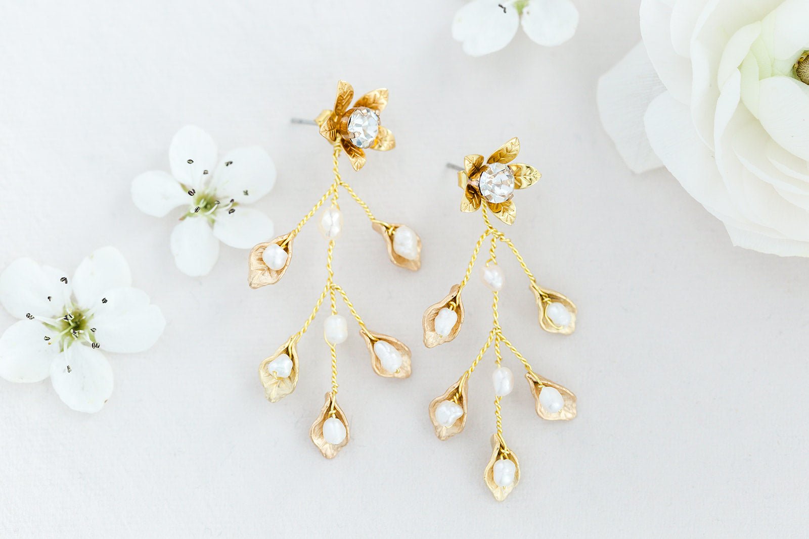 Gold floral bridal earrings with small fresh water pearl drops on a white background with white flowers. Statement bridal earrings handmade in Canada by Canadian jewelry maker