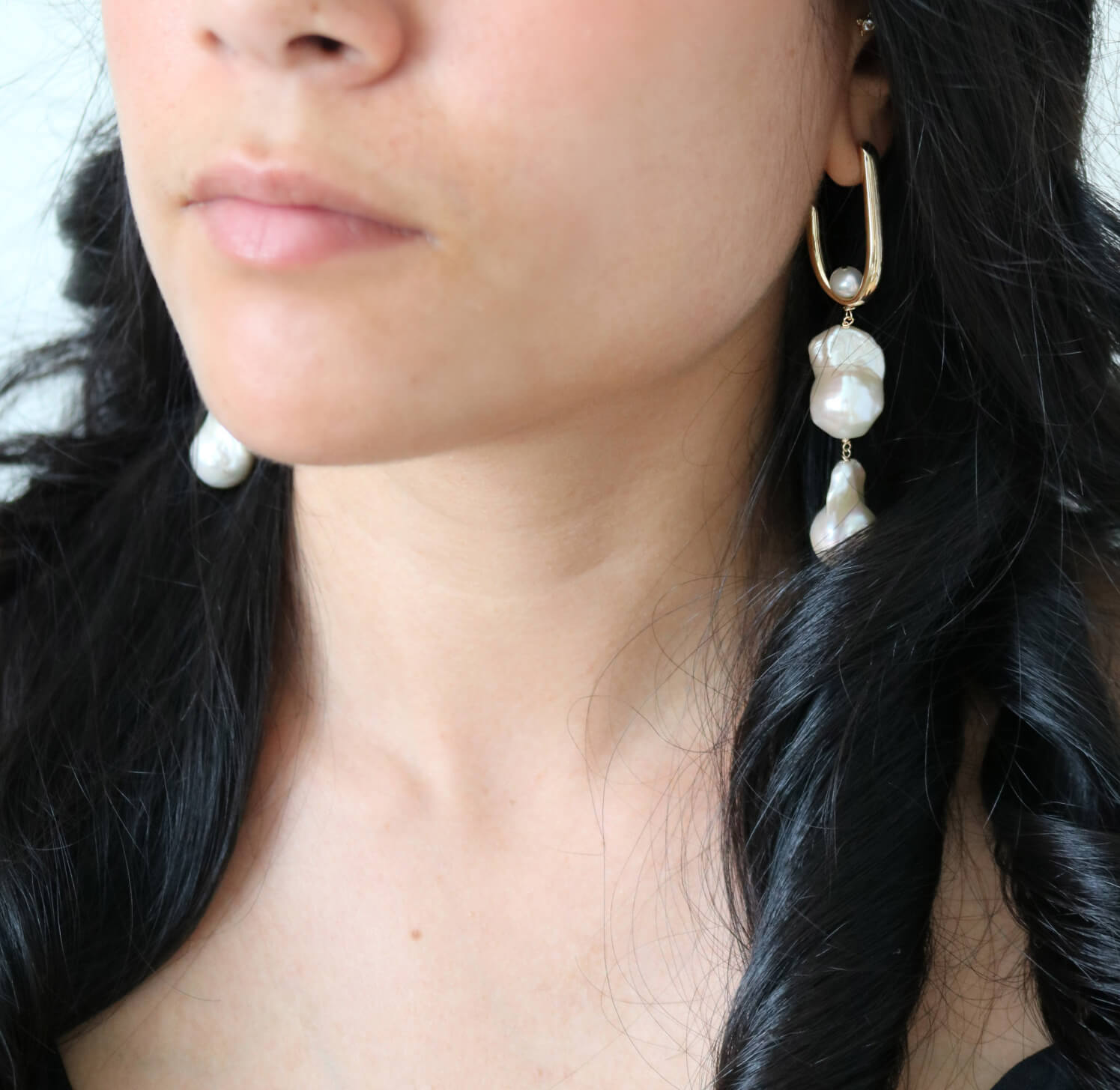 A close up of a dark haired woman wearing large statement bridal earrings in gold fill with two organic freshwater baroque pearls dangling down.