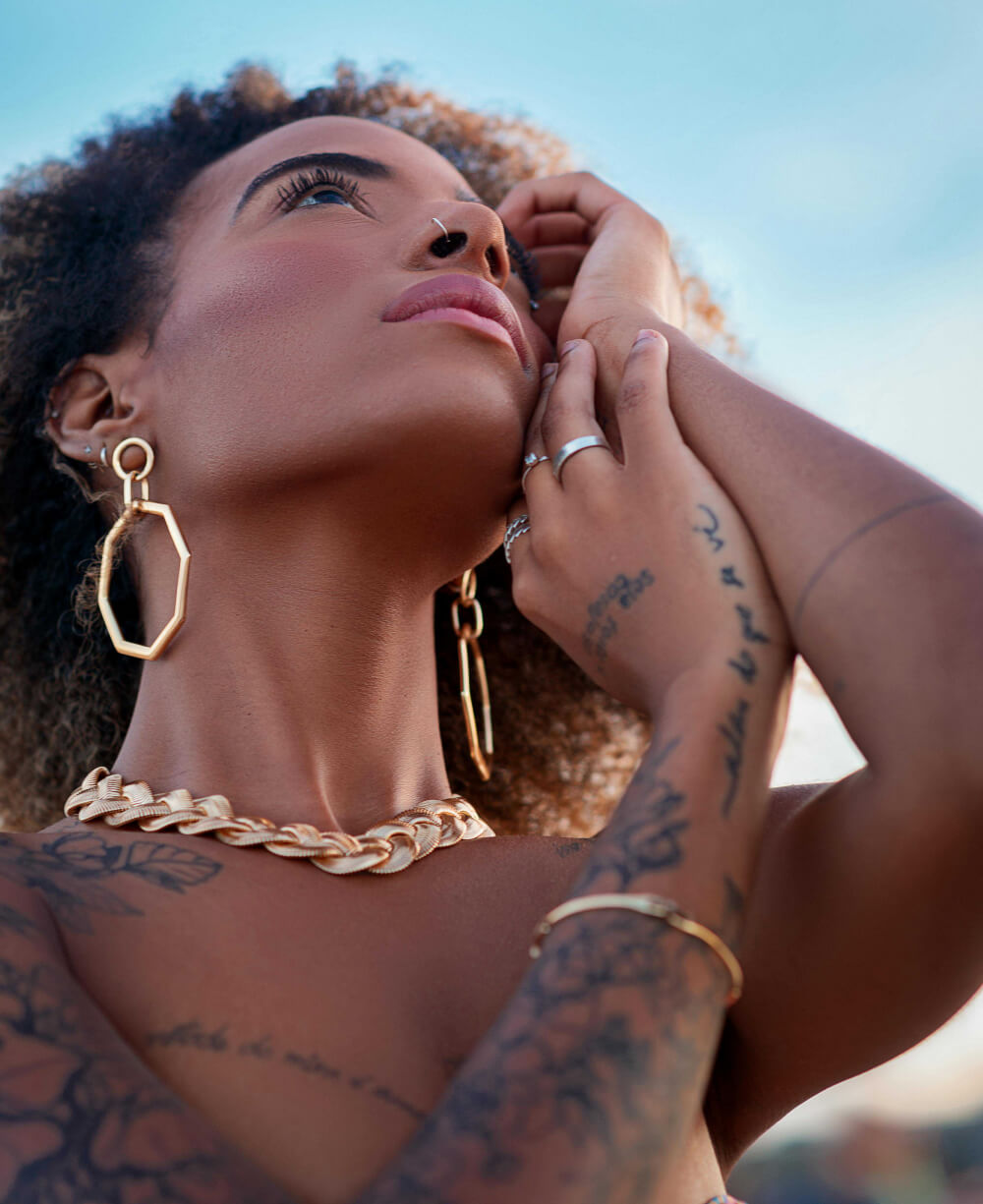A gorgeous BIPOC queen looking to the sky wearing gold statement jewelry with blue sky background.
