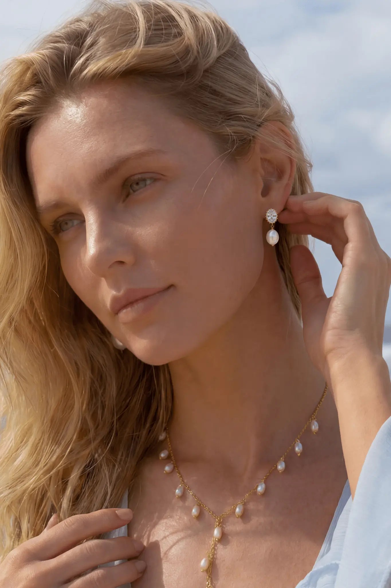 Close up of blonde woman on a beach wearing a light white dress, crystal pearl stud earrings, and gold chain lariat with freshwater pearl dangles. Canadian jewellery designs