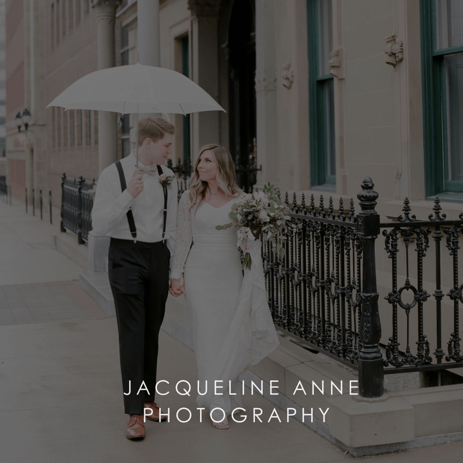 A newlywed couple walks the street hand-in-hand with a white umbrella and bridal flower bouquet.