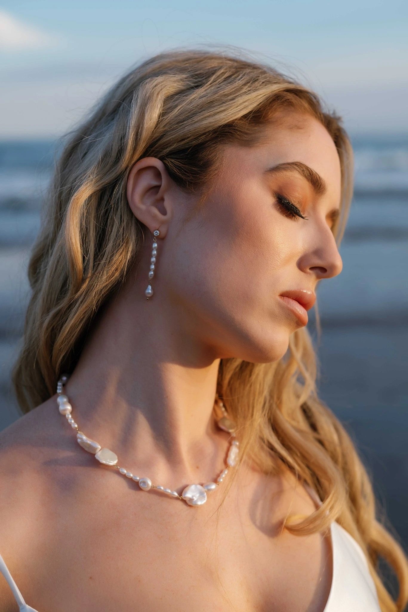 An ocean bride basks in the sun's rays while adorned in freshwater pearl dangle earrings and a multi-sized freshwater pearl necklace. Pearl jewelry sets & freshwater pearl bridal jewelry handmade by BIPOC wedding designer