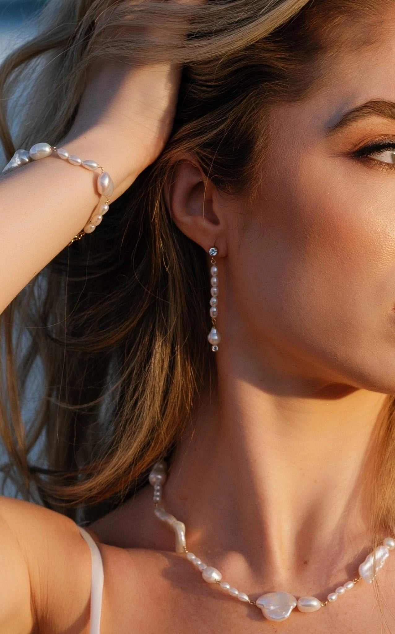 Close up of a bride on the beach at sunset with fingers in her hair wearing a freshwater pearl jewelry set with ocean background. Pearl jewelry sets & pearl bridal jewelry handmade by BIPOC jewelry designer