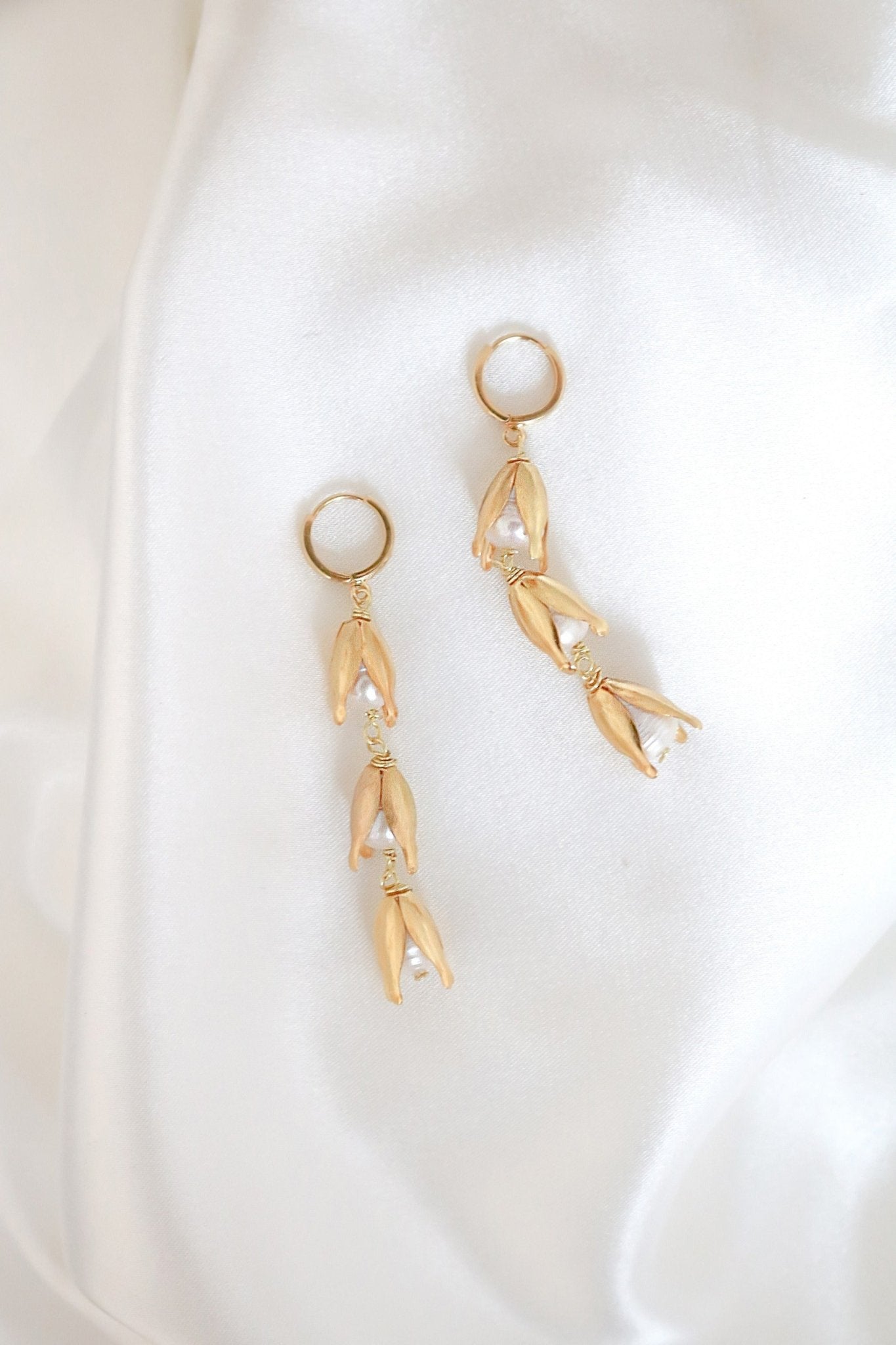 A gold-filled flower bud dangle earring with freshwater pearls on a huggie-style hoop lays on a sheet of white satin. Gold-filled jewelry Canada. Gold-filled Canadian jewelry. Canadian jewelry brands. Affordable Canadian jewelry brands. Handmade Canadian jewelry. Canadian handmade jewelry. Canadian bridal jewelry. Canadian jewelry business.