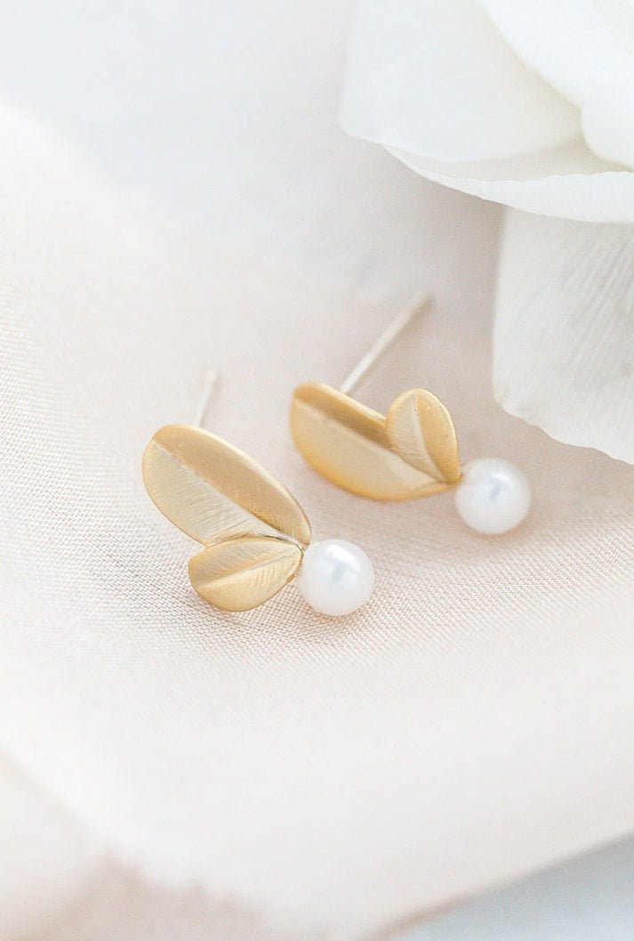 A pair of gold-filled leaf studs with sterling silver posts and freshwater pearls on a white linen background. Gold-filled jewelry