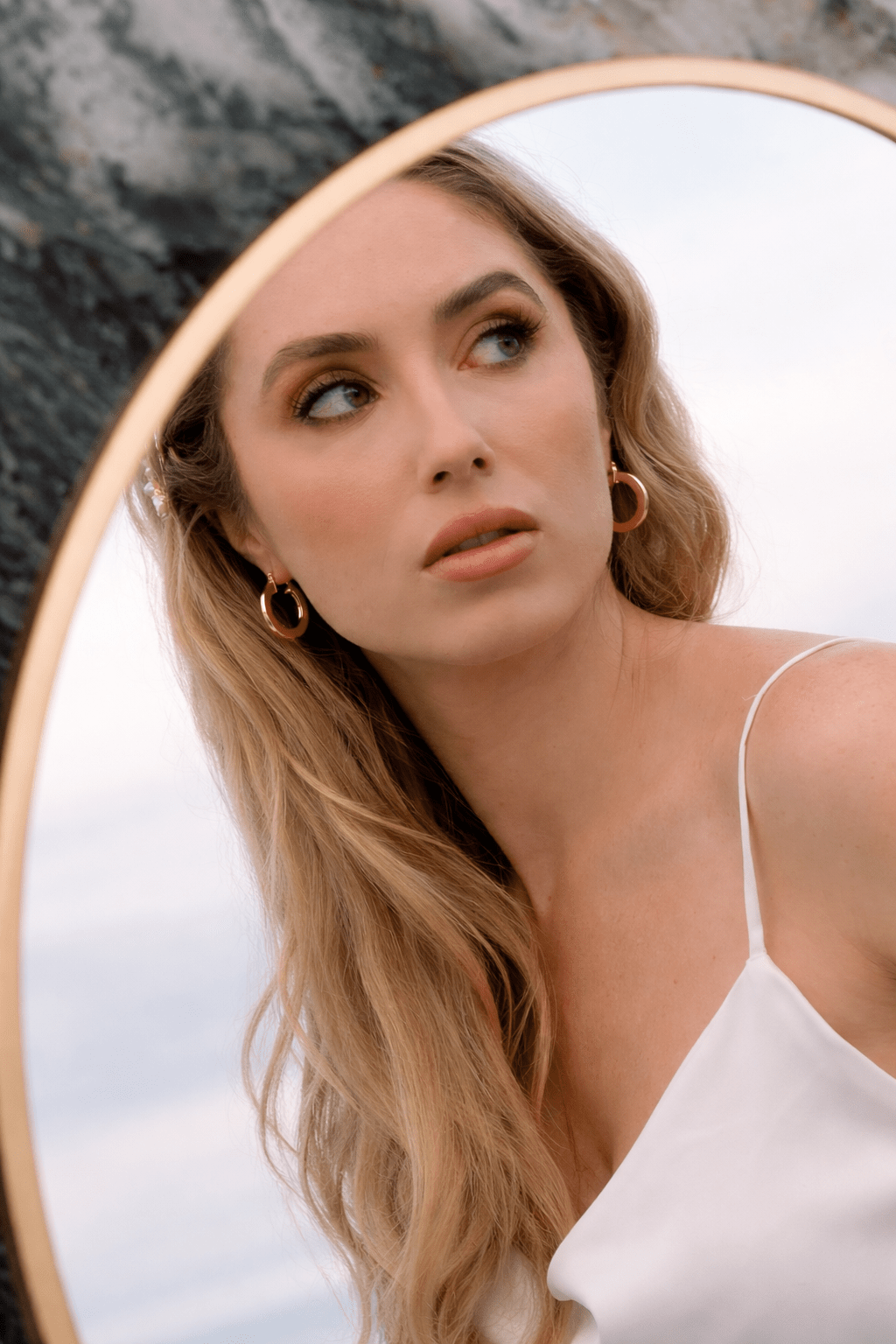 Bride with long blonde hair and a white bridal gown looking into a mirror with thick gold hoop earrings. Gold-filled Canadian jewelry, handmade modern hoops, & everyday earrings by BIPOC jewelry designer
