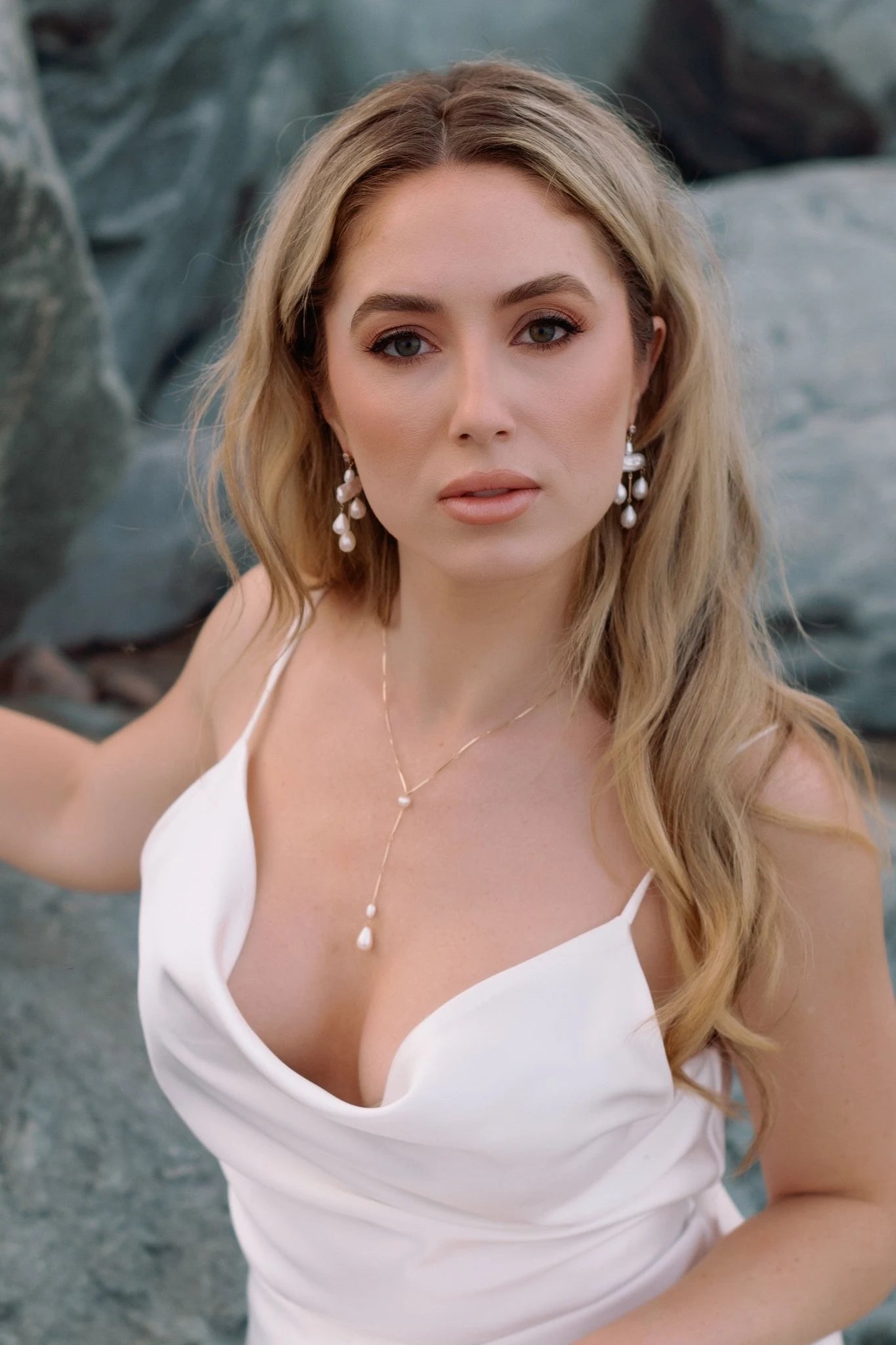 Beach bride in white wedding dress wearing a gold-filled lariat style pearl drop necklace & gold-filled freshwater pearl chandelier earrings with rocky background. Pearl jewelry sets, pearl bridal jewelry, & handmade bridal jewelry by Canadian bridal designer