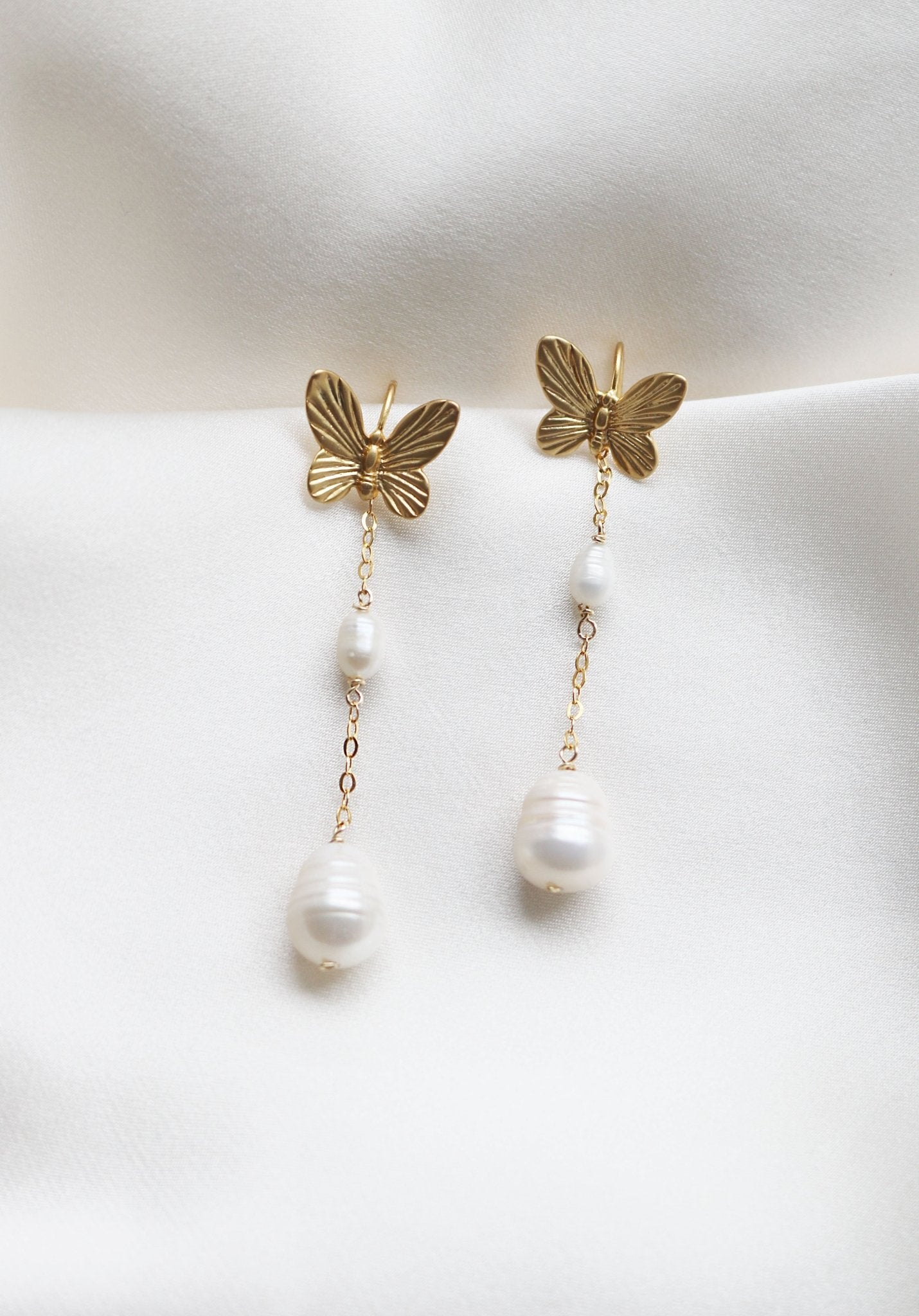 A pair of dangle Papillion Earrings in gold-filled with butterflies and freshwater pearls on a white satin sheet. Bridal jewelry Canada. Pearl jewelry Canada. Pearl Bridal jewelry Canada. Gold-filled jewelry Canada. Canadian bridal jewelry. Woman-owned Canadian jewelry brand. Canadian jewelry business. Organic pearl jewelry.