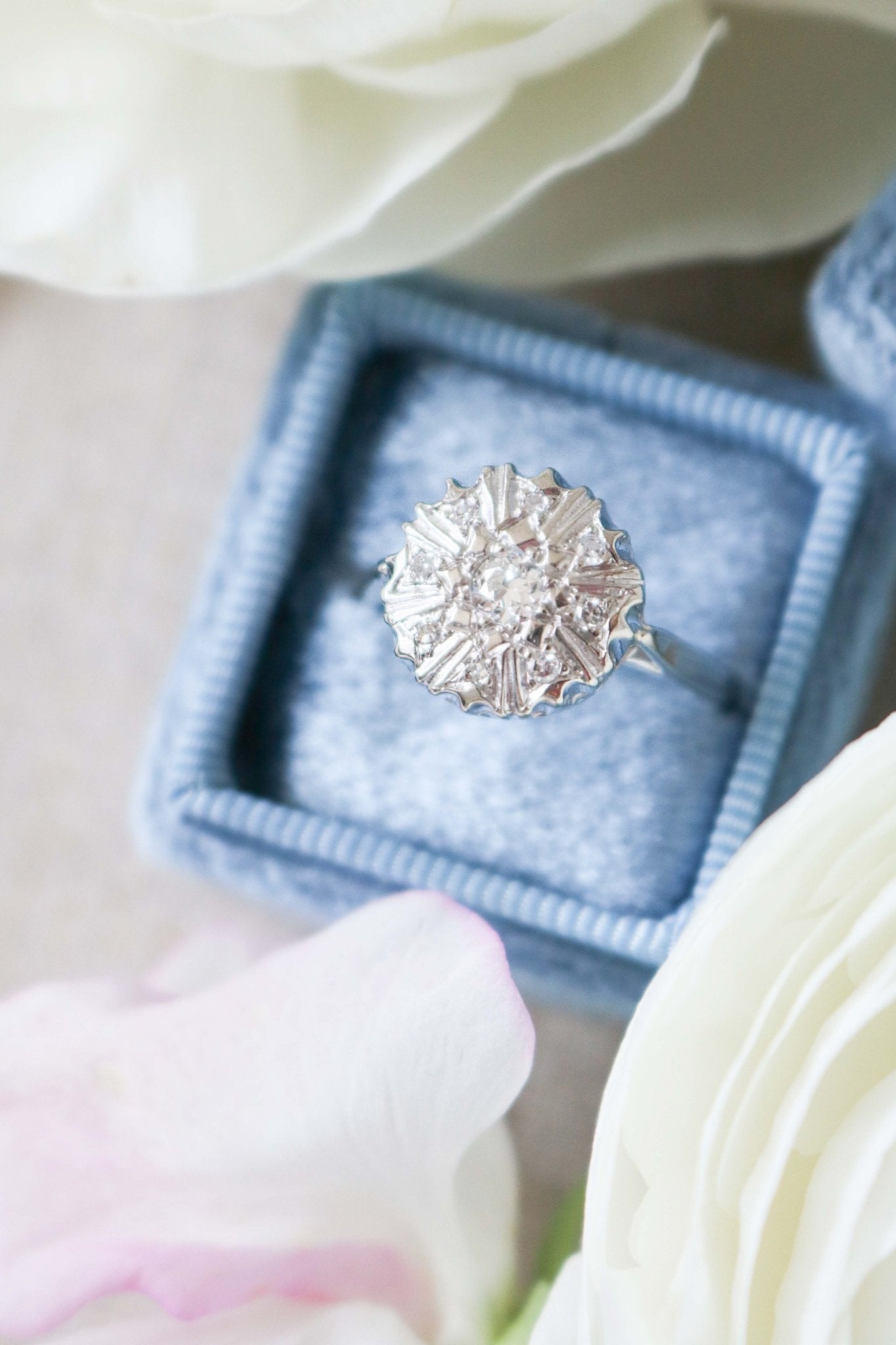 An extreme close up of top view of Selene Art Deco 14k White Gold Diamond Vintage Engagement Ring in a blue ring box surrounded by white roses. Canadian vintage engagement rings. Bridal jewelry Canada. Vintage engagement rings Canada. Canadian weddings. Wedding Canada. Woman-owned Canadian jewelry brand. Canadian jewelry business. Small Canadian jewelry brands.
