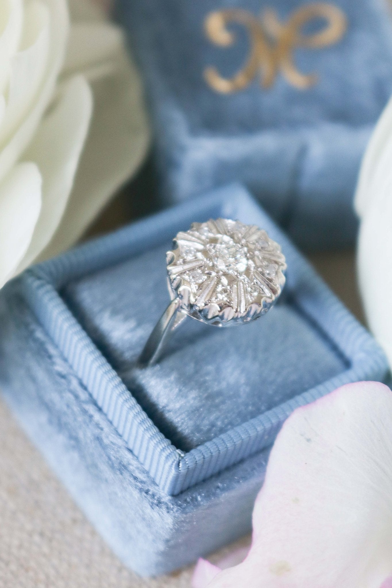 An extreme close up side view of the Selene Art Deco 14k White Gold Diamond Vintage Engagement Ring in a blue ring box surrounded by white roses. Canadian vintage engagement rings. Bridal jewelry Canada. Vintage engagement rings Canada. Canadian weddings. Wedding Canada. Woman-owned Canadian jewelry brand. Canadian jewelry business. Small Canadian jewelry brands.