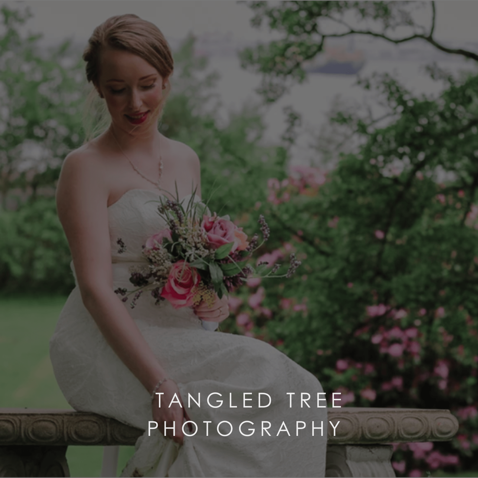 A bride in white sits on a marble bench in spring with a small bouquet of pink roses.