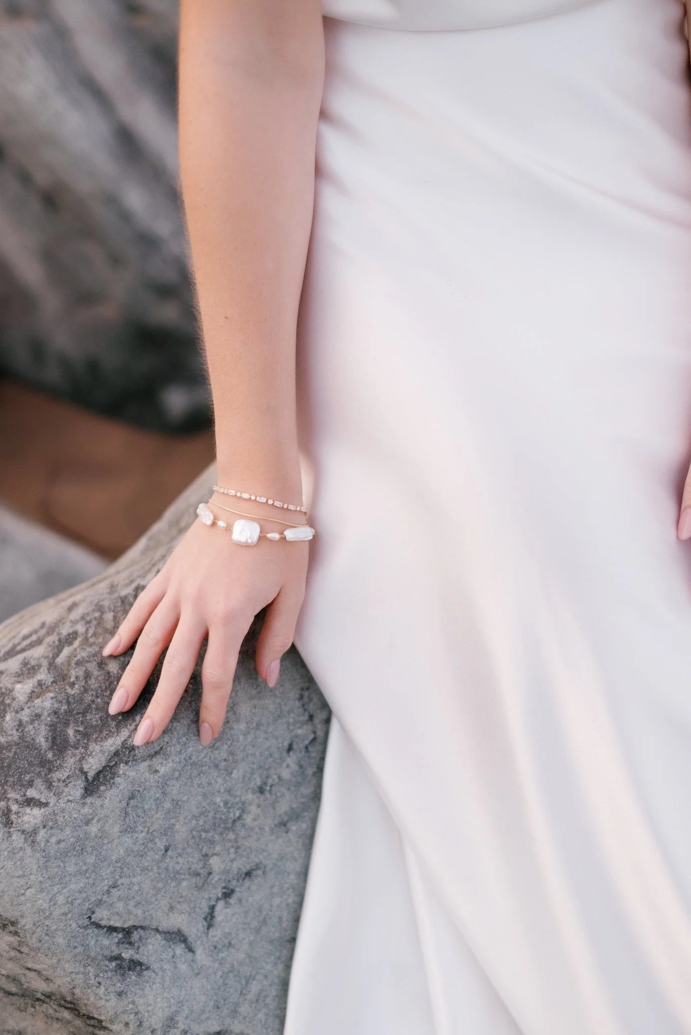 A bride sits on a beach rock in a sleek wedding gown. She wears three bracelets stacked for heightened allure; gold filled crystal, simple strand, and thick freshwater pearl. Pearl Bridal jewelry handmade in Canada