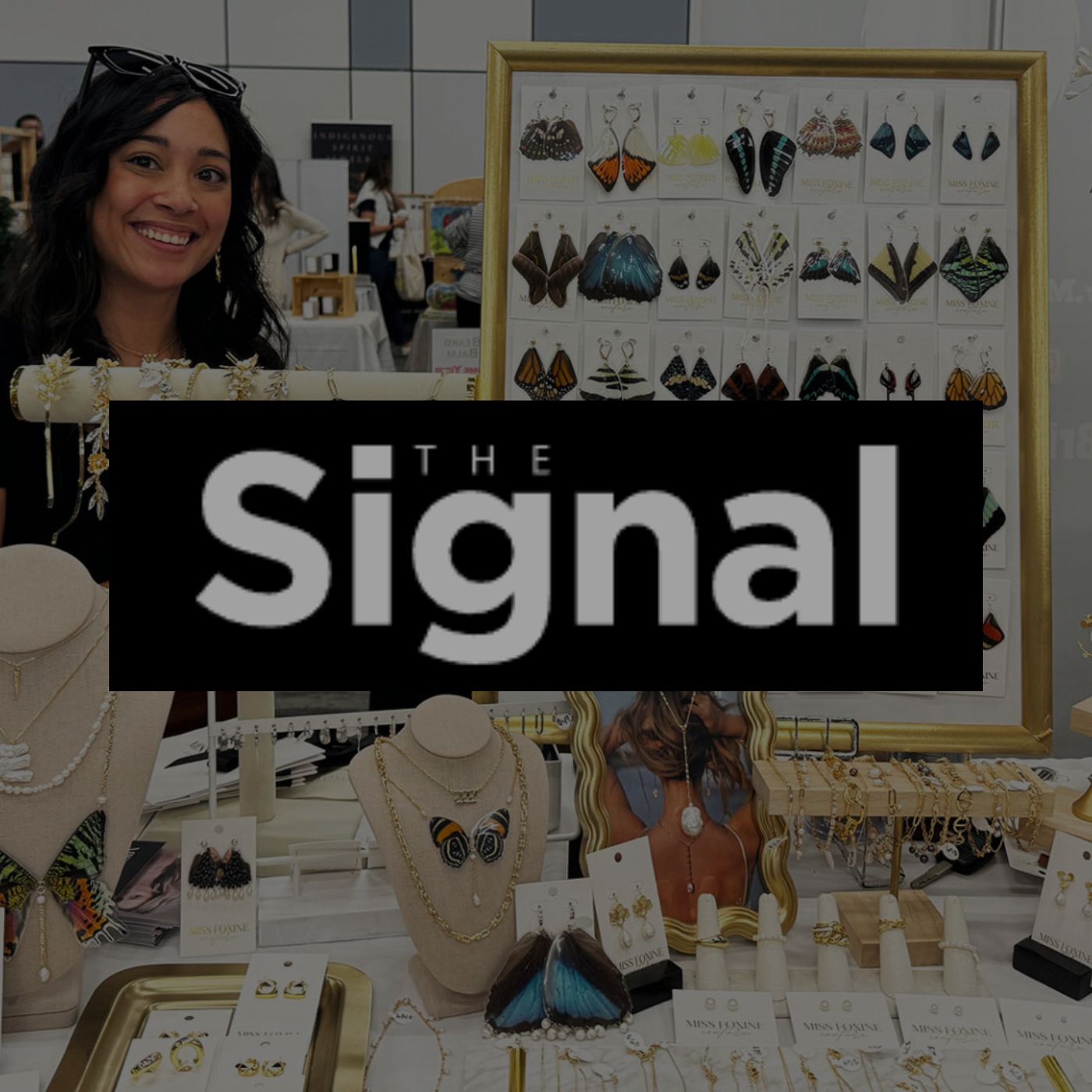 A black and white logo for The Signal article with background of Miss Foxine Couture display featuring butterfly wing earrings, gold-filled bracelets, rings, and freshwater pearl necklaces.