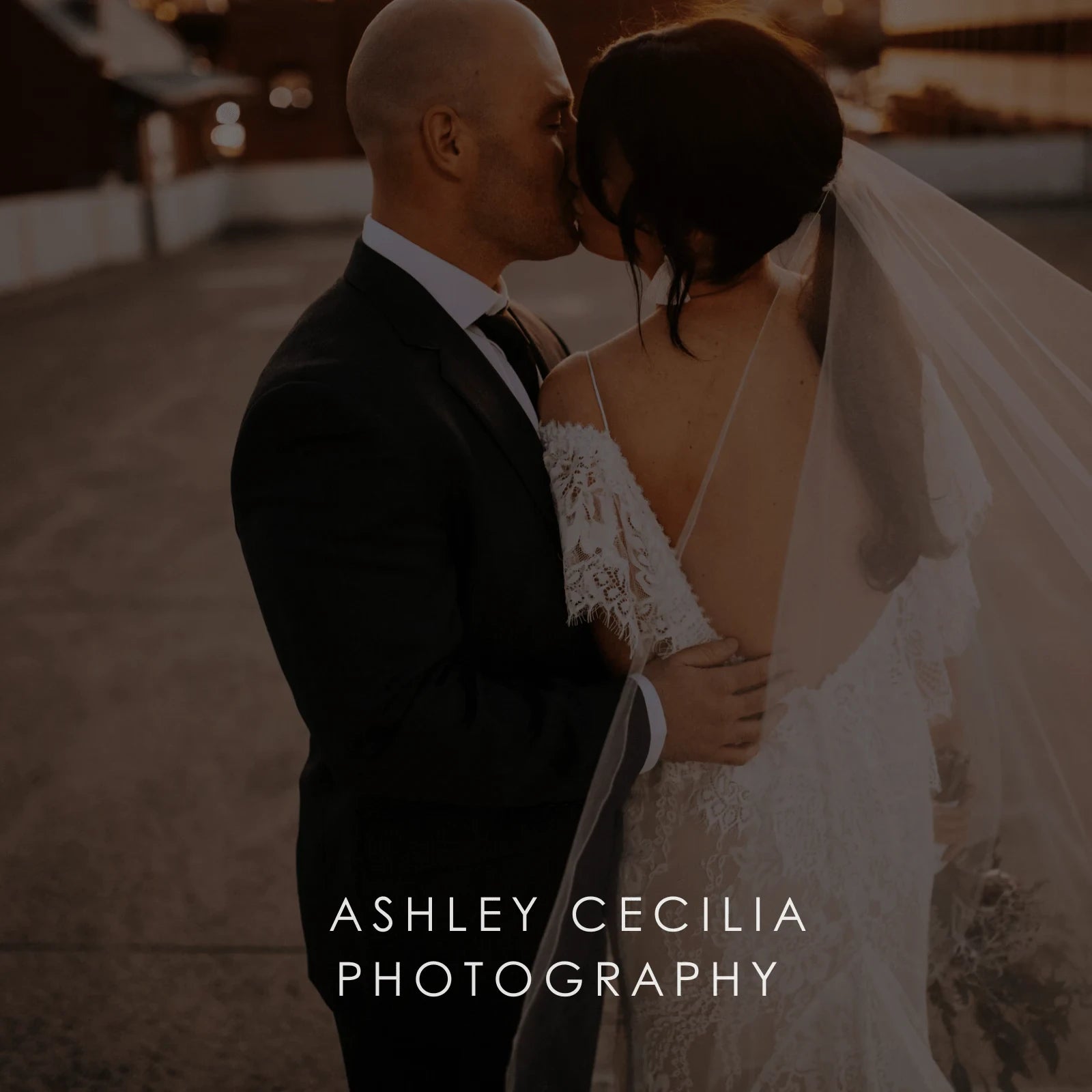 A bride in wedding gown and long tulle veil and groom kiss at sunset.
