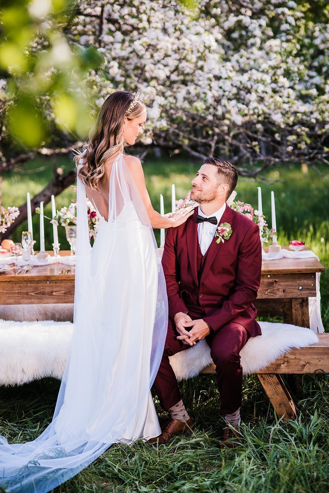 A wedding veil cape flows behind a bride as she touches her groom's shoulder while he's sitting at a decorated picnic table under a flowering tree. Wedding veils and bridal veils handmade in Canada for weddings by Canadian wedding designer