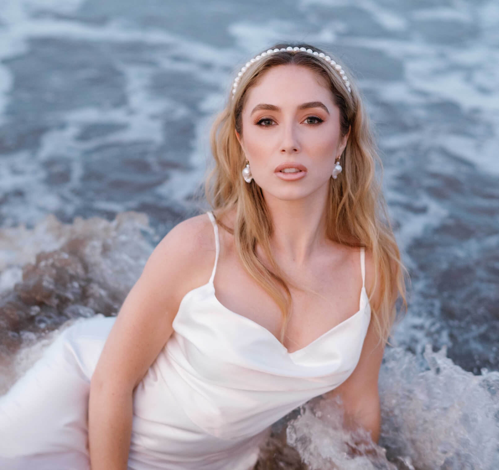 A bride lays in the ocean waves in her sleek white wedding dress and freshwater pearl bridal jewelry.