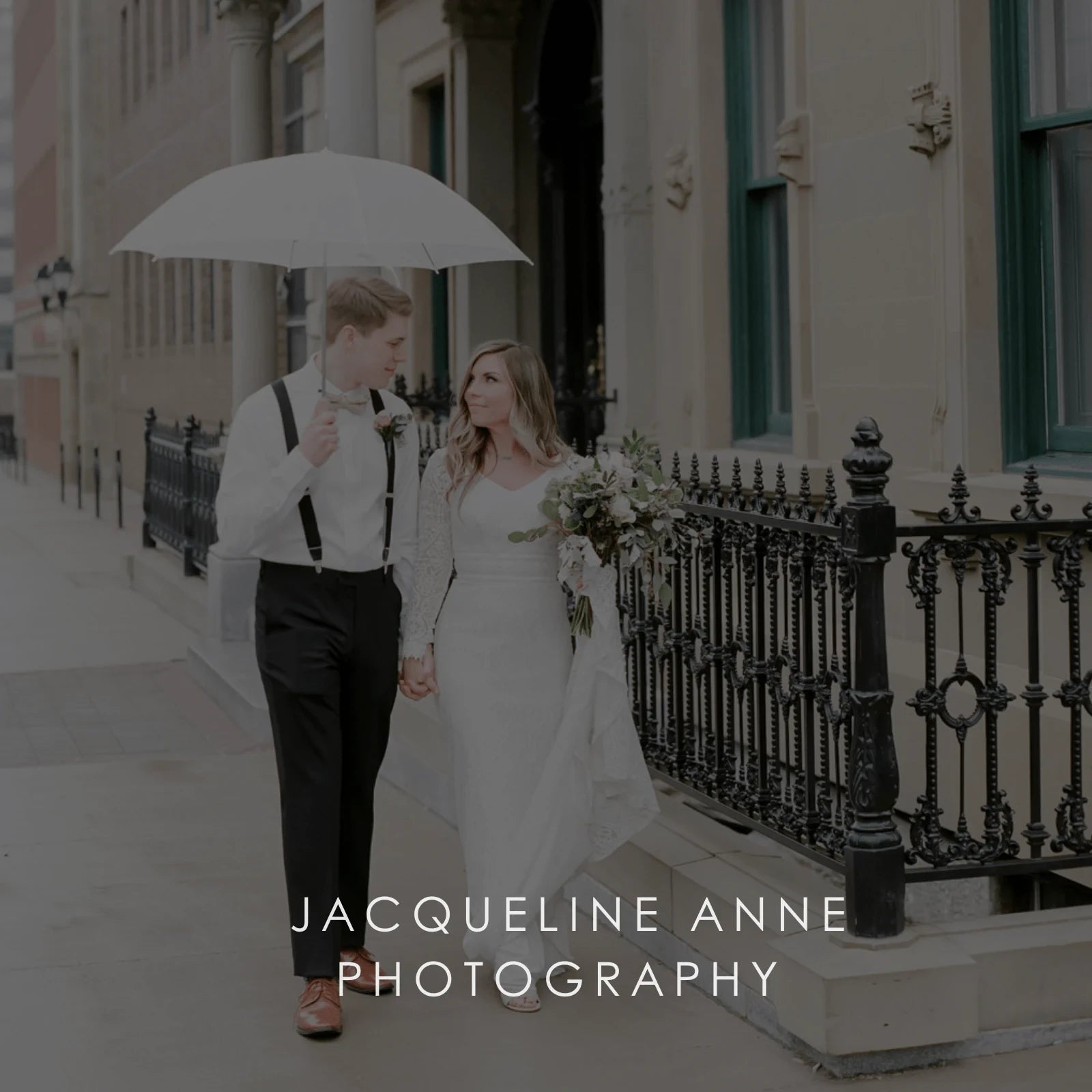 A newlywed couple walks the street hand-in-hand with a white umbrella and bridal flower bouquet.