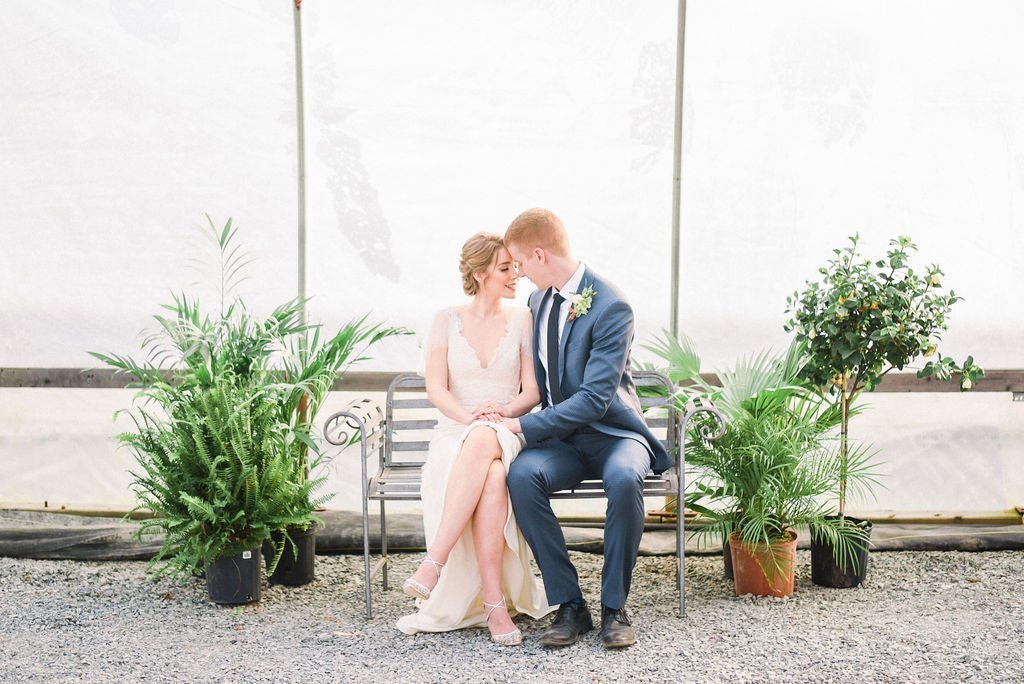A bride and groom sit together on a bench in a greenhouse.