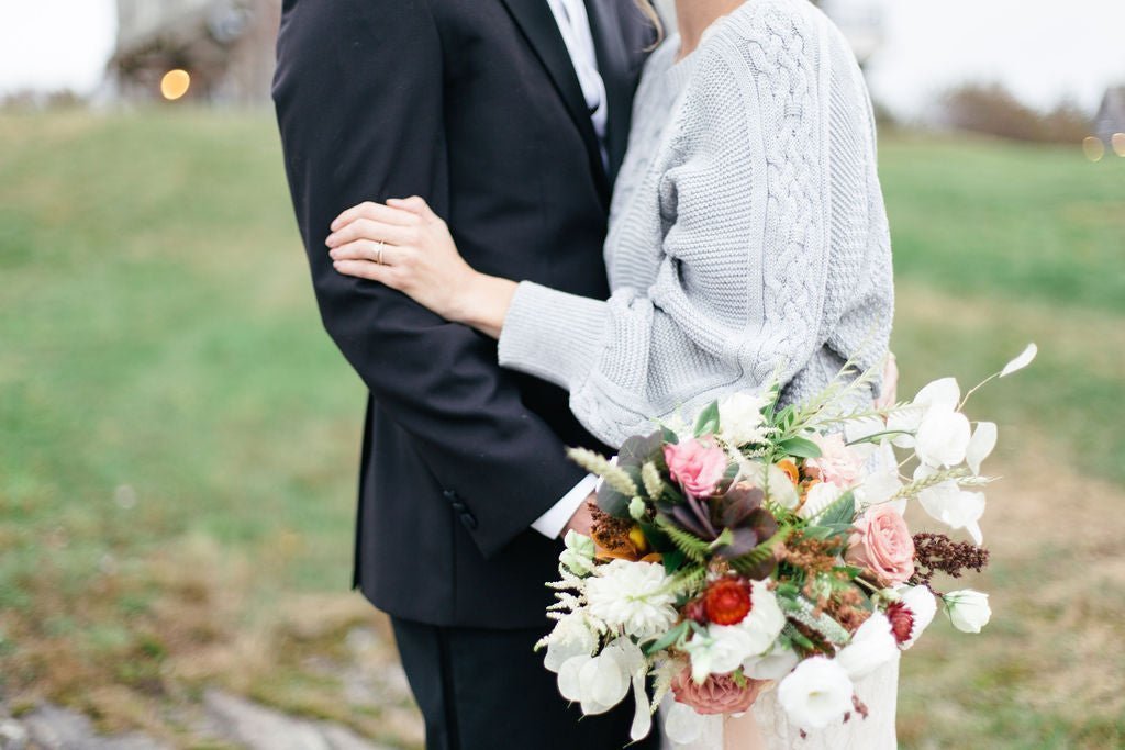 A cozy bride and groom embrace while holding a bridal flower bouquet.