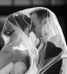 A black and white photo of a bride and groom touching foreheads shrouded by a white tulle wedding veil