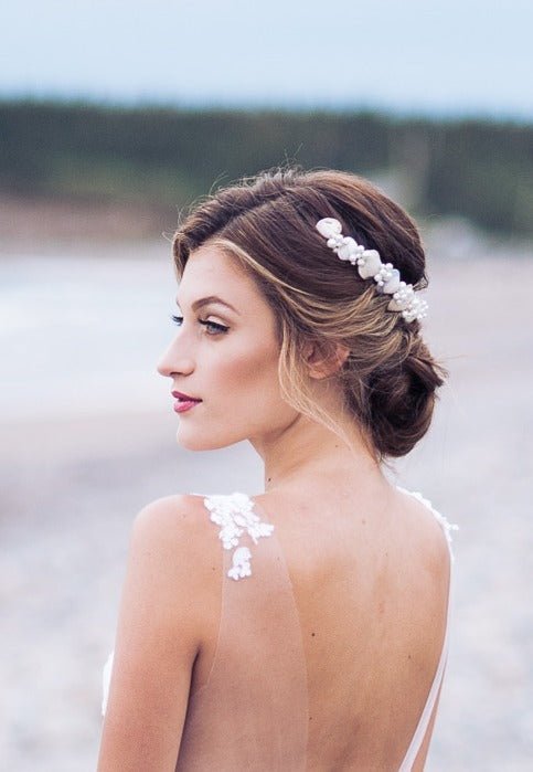 Bride in a sheer lace wedding gown looks out to sea while wearing a seashell and freshwater pearl hair wreath. 