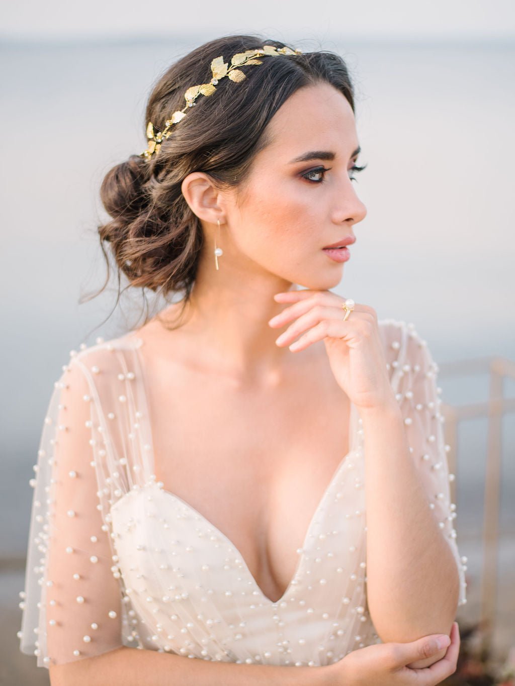 Thoughtful bride contemplates while displaying a beautiful golden hair circlet with dainty freshwater pearls and crystals among glistening leaves. 