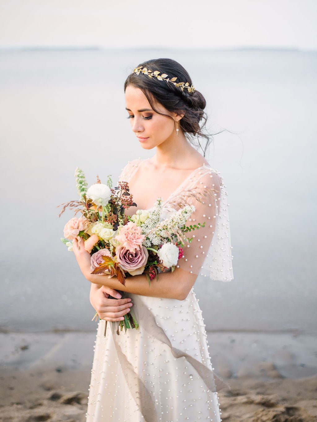 Beautiful bride in pearl-dotted wedding dress holds flower bouquet while wearing a golden circlet with leaves, pearls, and crystals. 