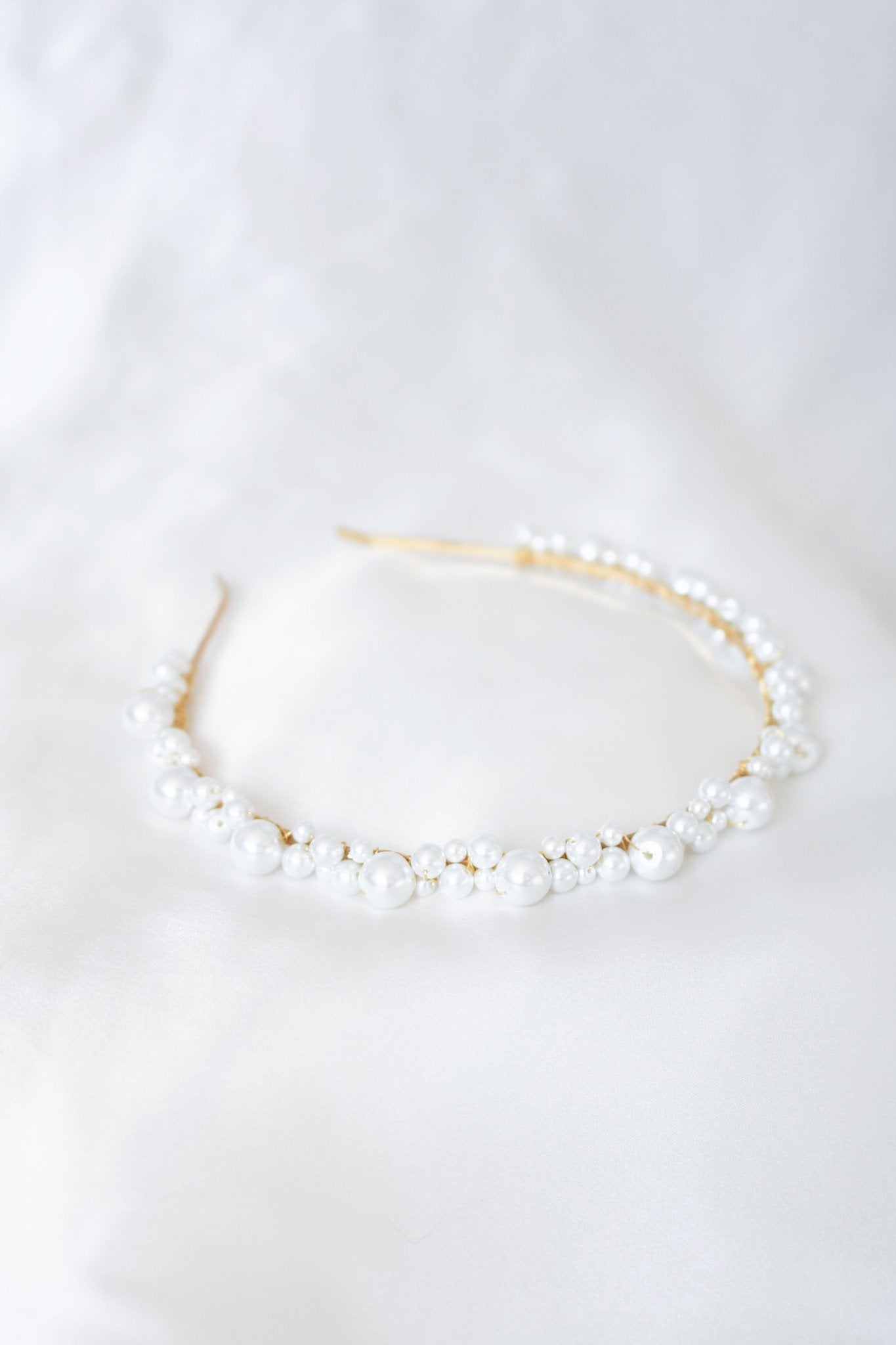 A golden wire-wrapped headband coated in pearls lays on a white satin sheet. 