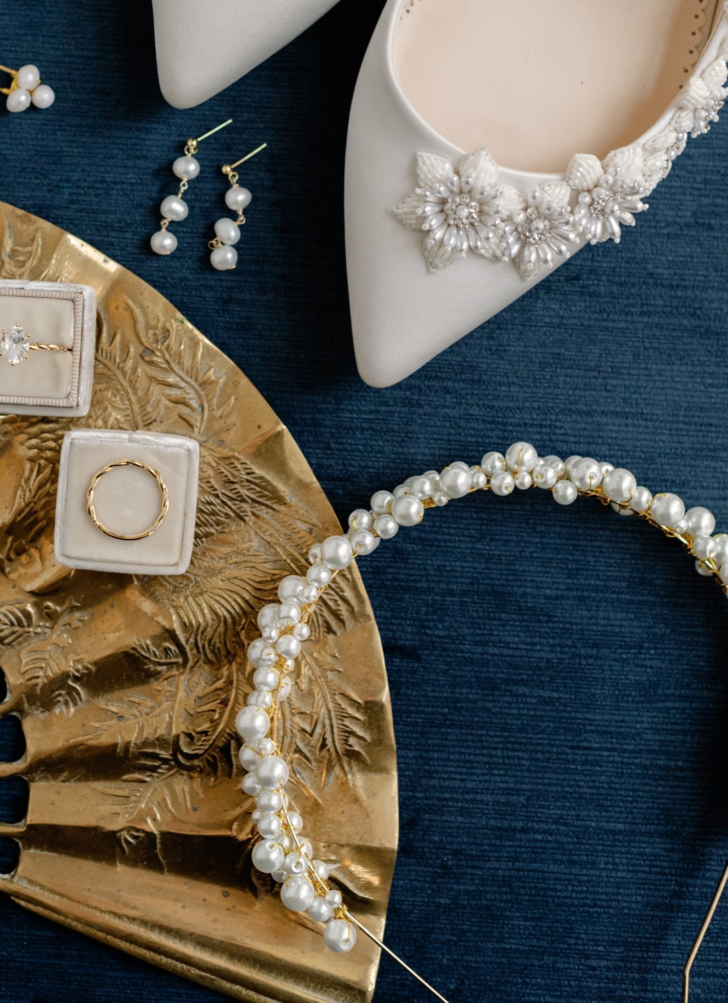 A gold bridal pearl headband with clustered Swarovski pearls displayed with decorated bridal shoes, Pearl Dangle Earrings, and Gold Rings on a blue fabric surface.