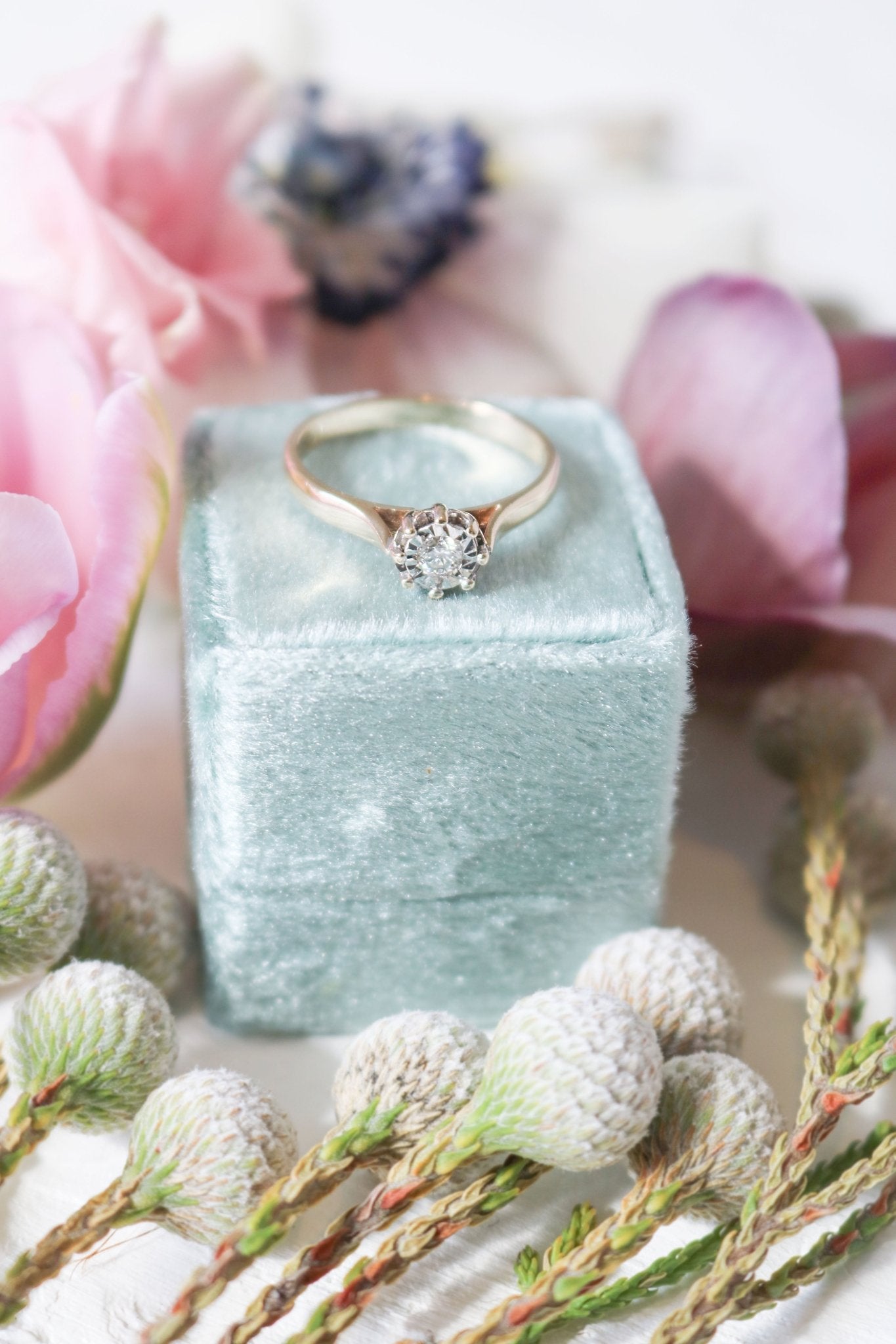 An Antique Vintage Engagement Ring in 9ct Yellow and White Gold with Diamond on a ring box among flowers.