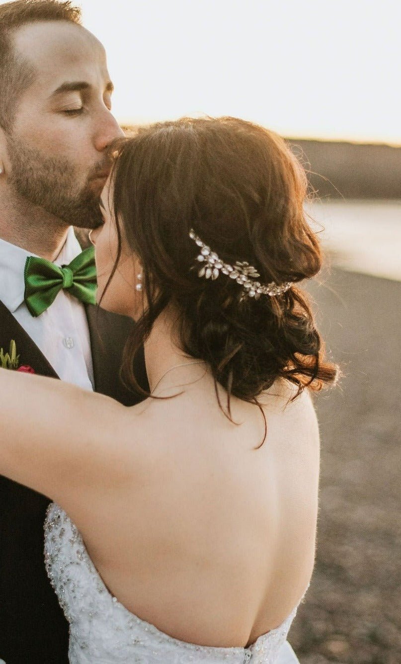 A groom kisses his bride's forehead. Her silver crystal bridal hair crown accentuates her dark curly hair with crystals. 