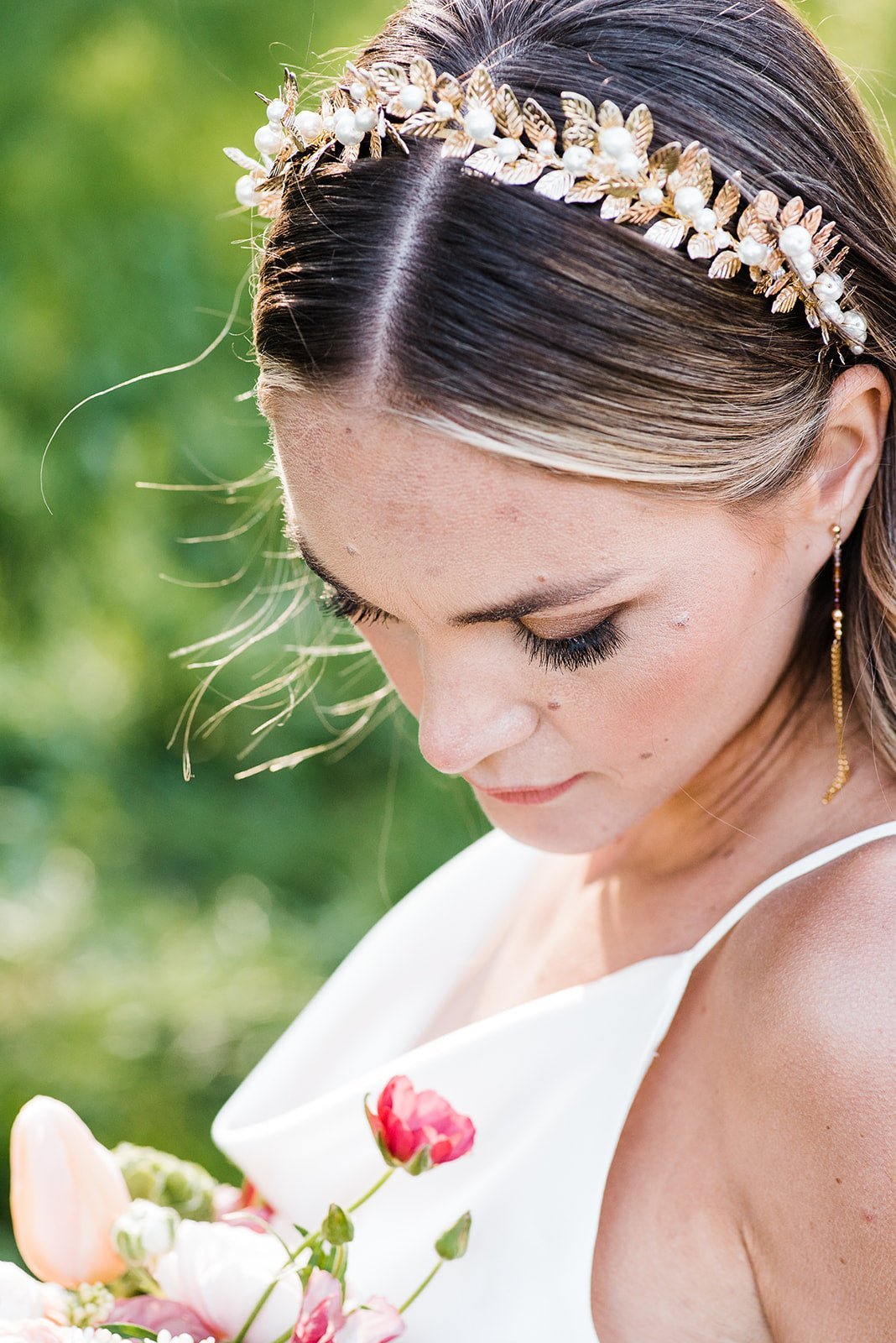 A bride gazes down at some flowers while showcasing a large gold and pearl bridal headband.