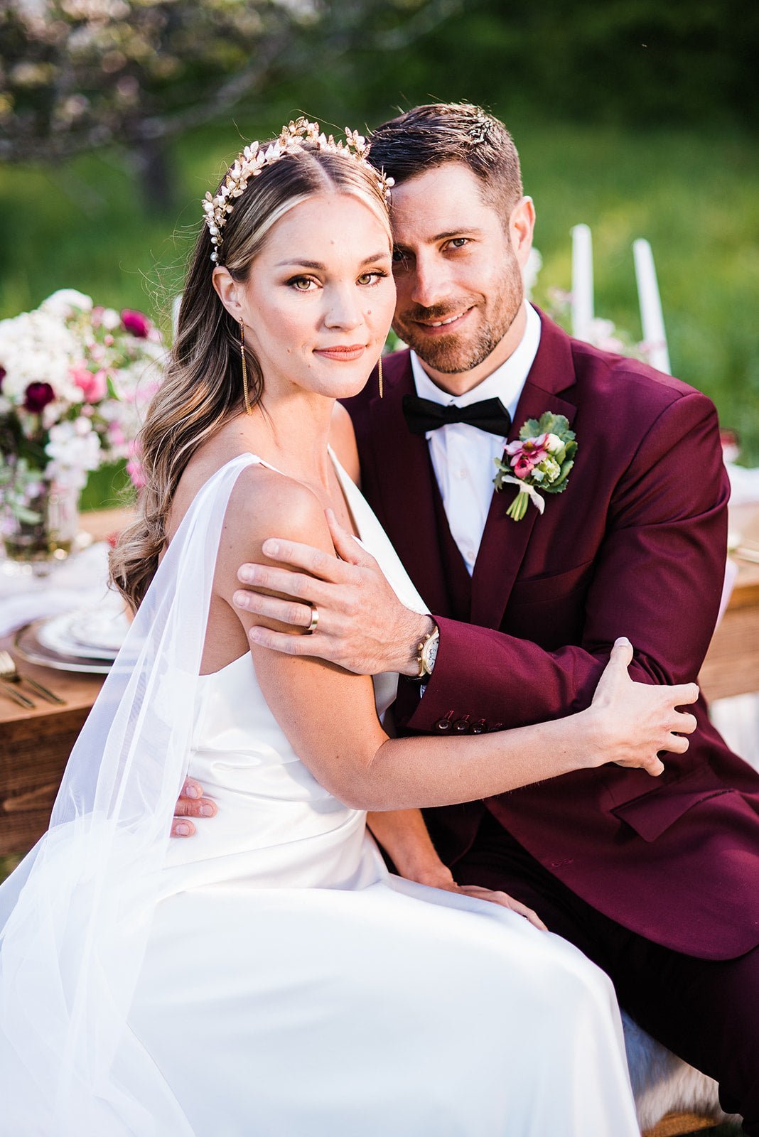 A beautiful newly married couple sit together at an outdoor table. She looks like a goddess in her Artemis laurel leaf/pearl crown.